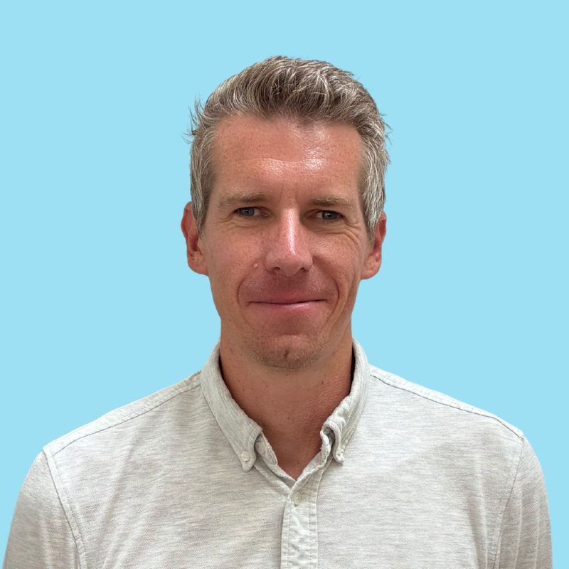 Portrait of a man with short light brown hair, glasses, and a slight smile, wearing a collared shirt and a dark jacket against a light blue background.