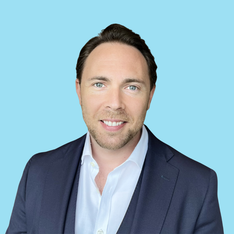A smiling man with dark hair, wearing a beige blazer and a white shirt, standing against a light blue background.