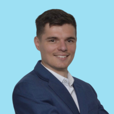 A man with short brown hair, wearing a blue suit, smiling against a light blue background.