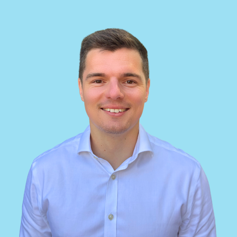 A man with short brown hair, wearing a blue suit, smiling against a light blue background.