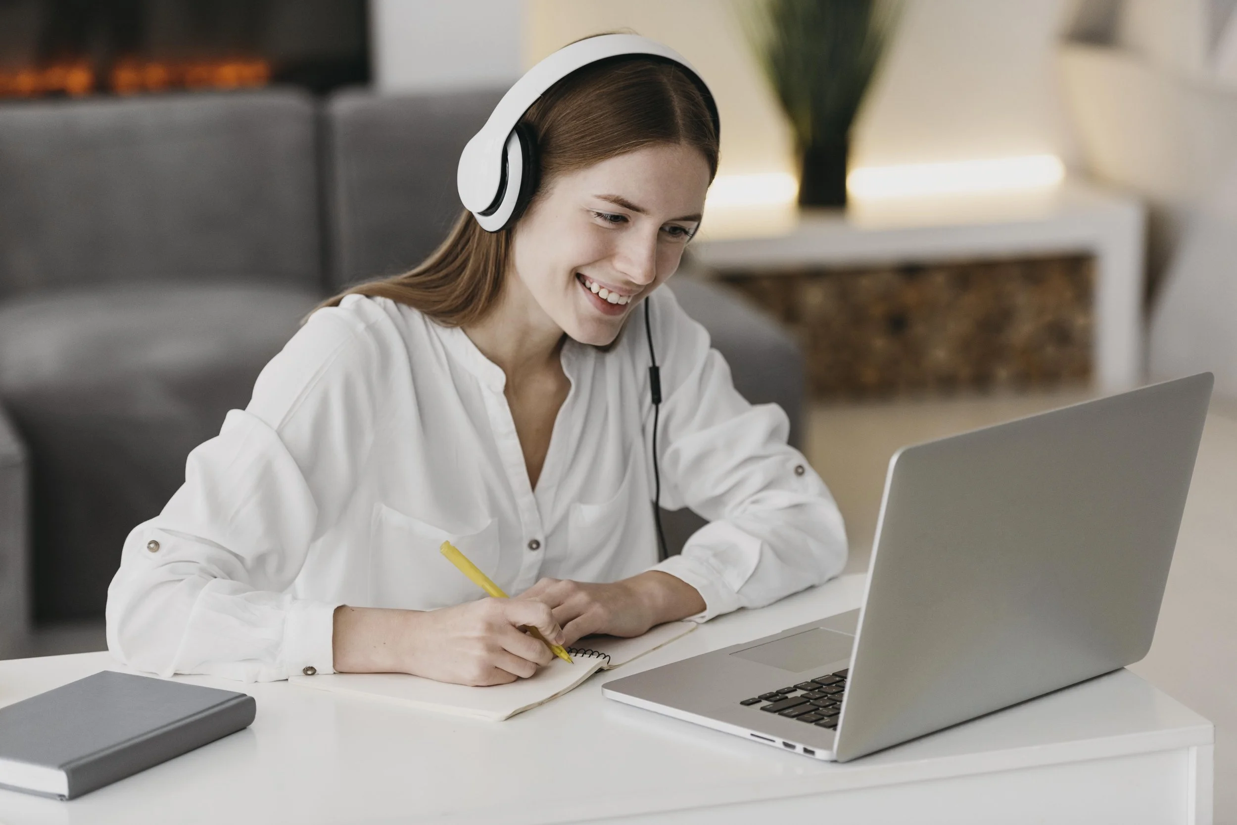 A woman with long brown hair wearing a white blouse and headphones, sitting at a desk with a silver laptop, writing in a notebook, smiling while looking at the laptop screen.