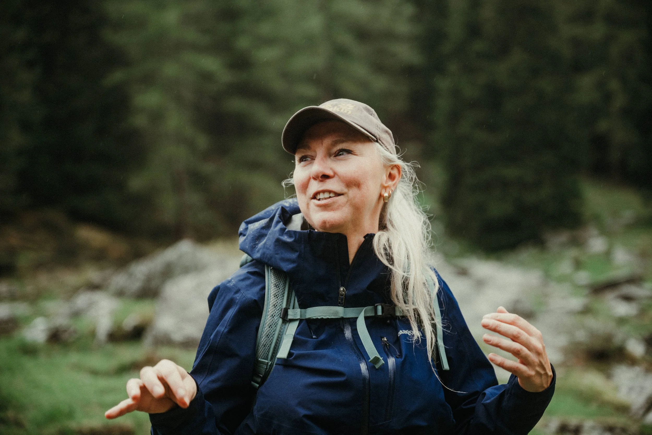 An elderly woman with gray hair and sunglasses standing on large rocks in a rocky outdoor landscape, with two other people partially visible in the foreground.