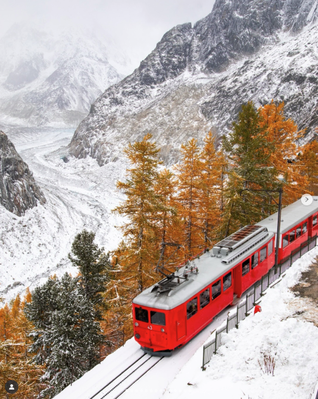 A red train traveling through a snowy mountain landscape with colorful autumn trees and snow-covered mountains in the background.