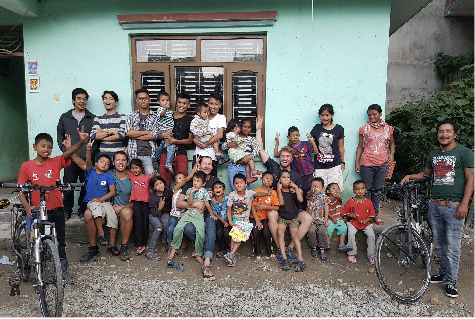 A large group of children and adults gathered outdoors in front of a teal building, smiling and posing for a photo, with two bicycles at either side of the group.
