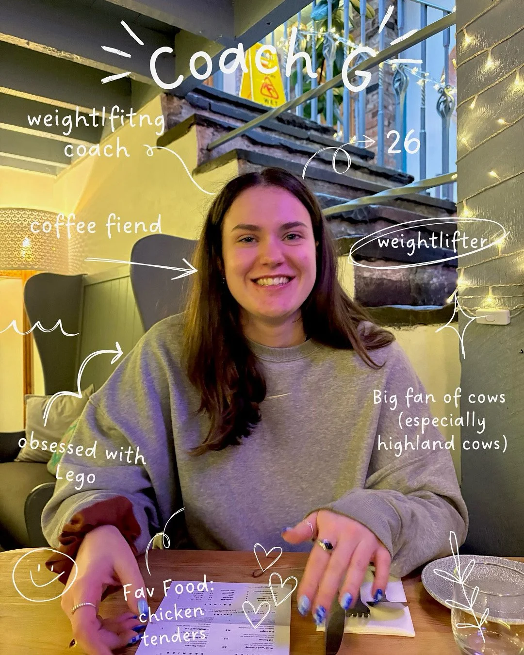 A young woman with brown hair smiling at a restaurant table. She is wearing a grey sweatshirt and has painted nails. The background shows a staircase and brick wall. Hand-drawn annotations describe her interests, age, job, and favorite food, mentioning she is a coach, a weightlifter, and a fan of highland cows, especially their horns.