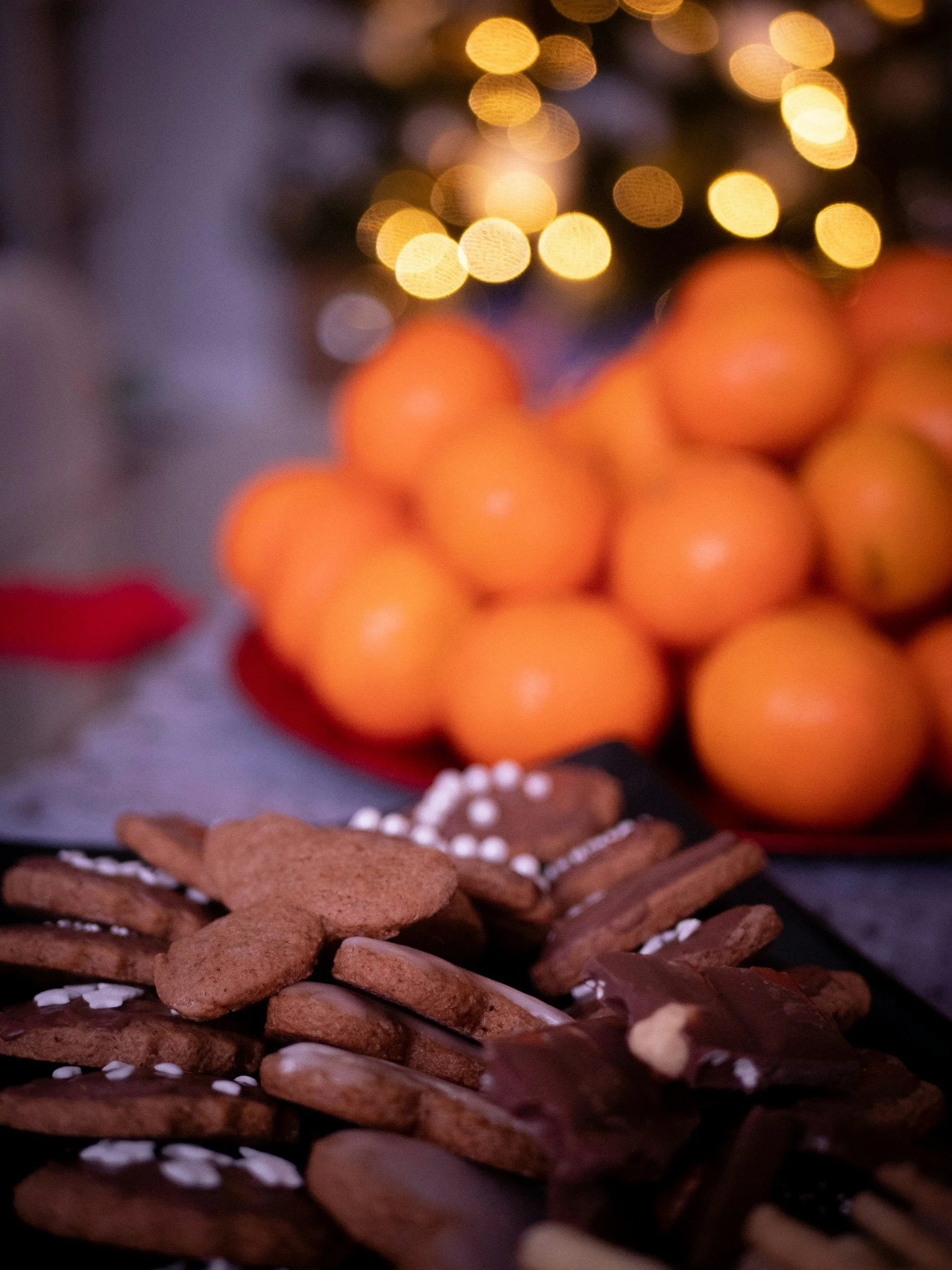Close-up of assorted Christmas cookies in front of a blurred Christmas tree with warm lights, and oranges in the background.