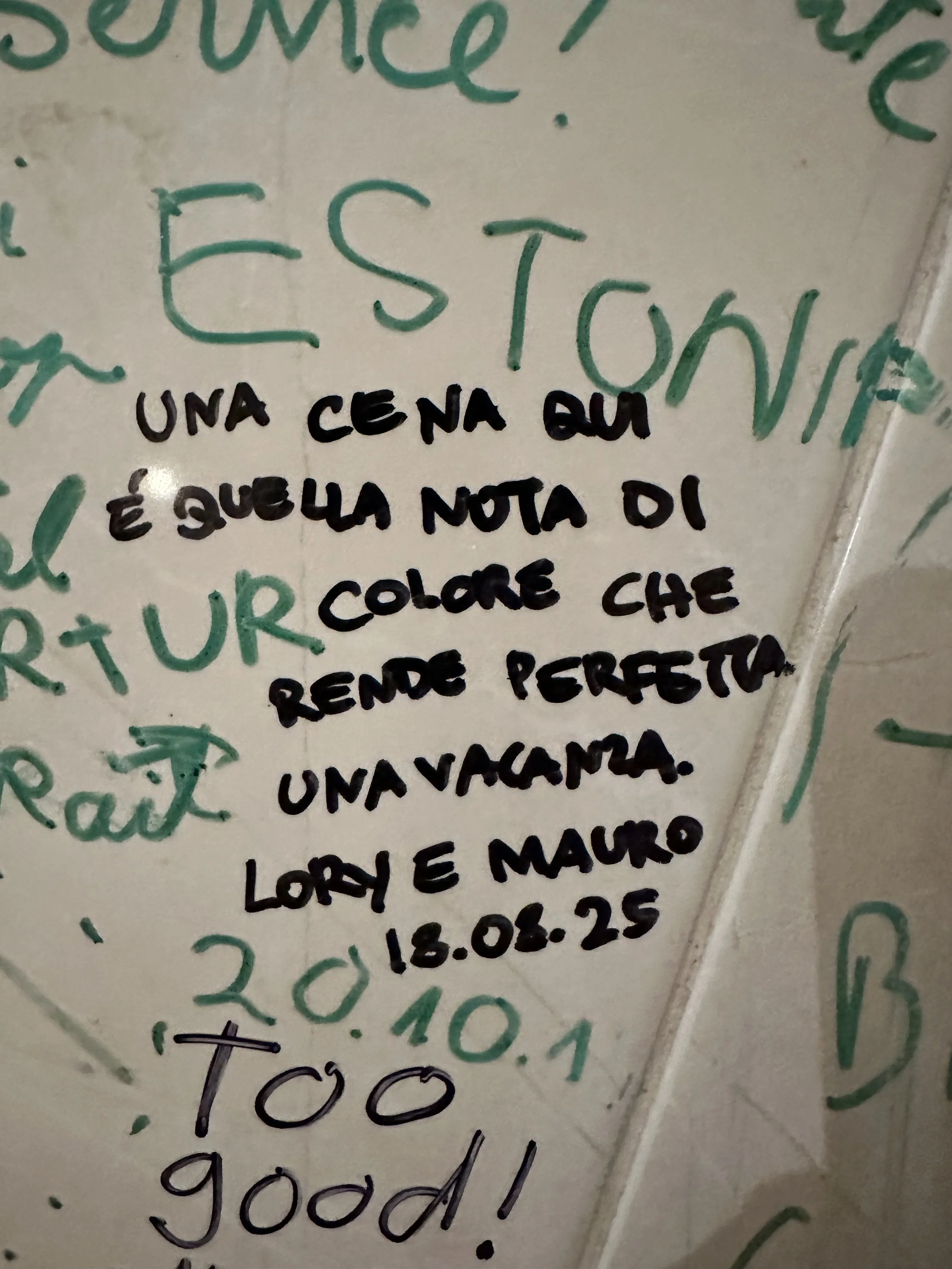 Italian handwritten note on a wall describing a dinner as a note of the color that makes the sunset a vacation. Signed by Lori and Mauro, dated 18.08.25, with additional notes and text around.
