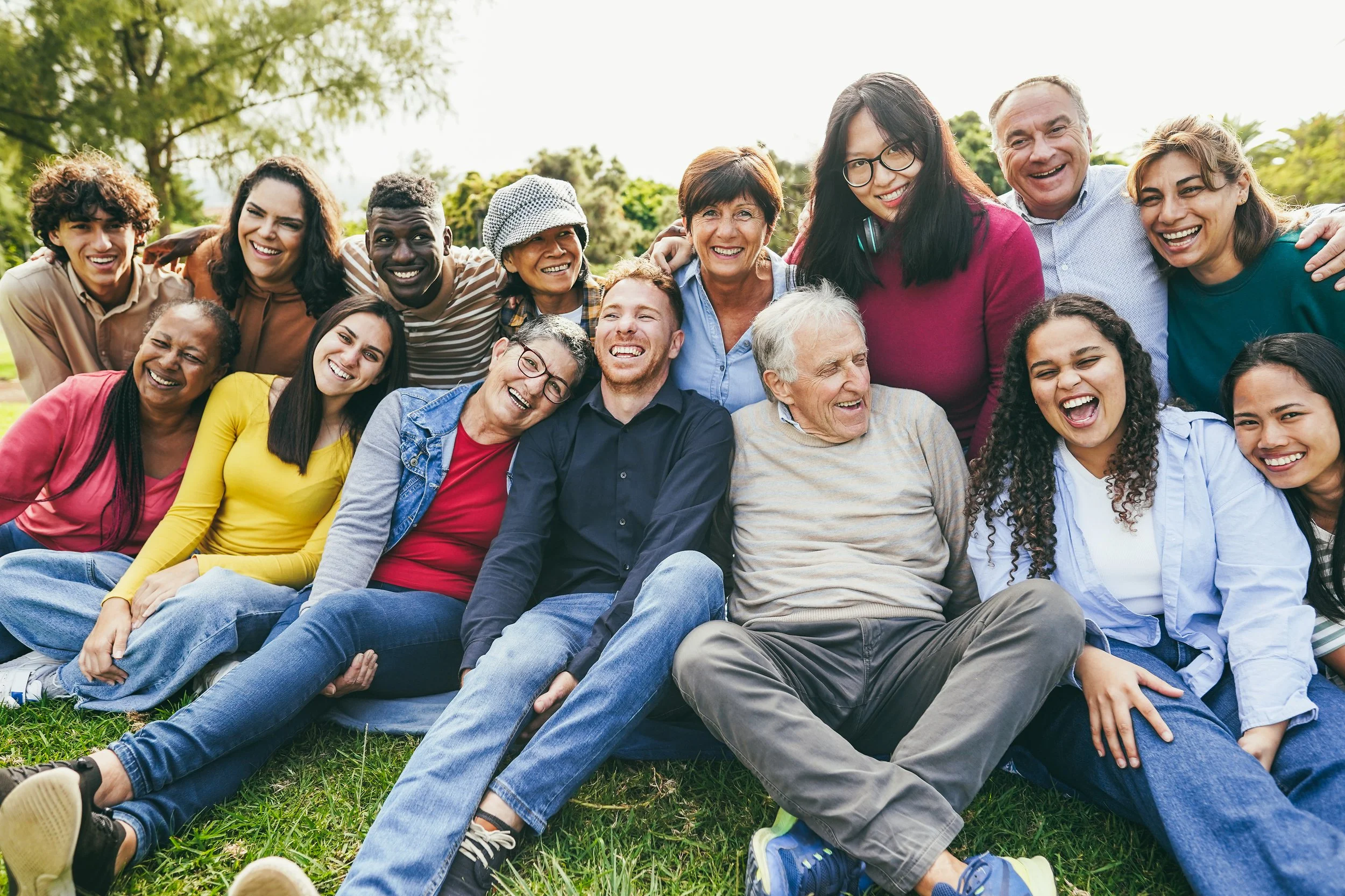 Group of diverse people sitting and standing outdoors on green grass, smiling and laughing.
