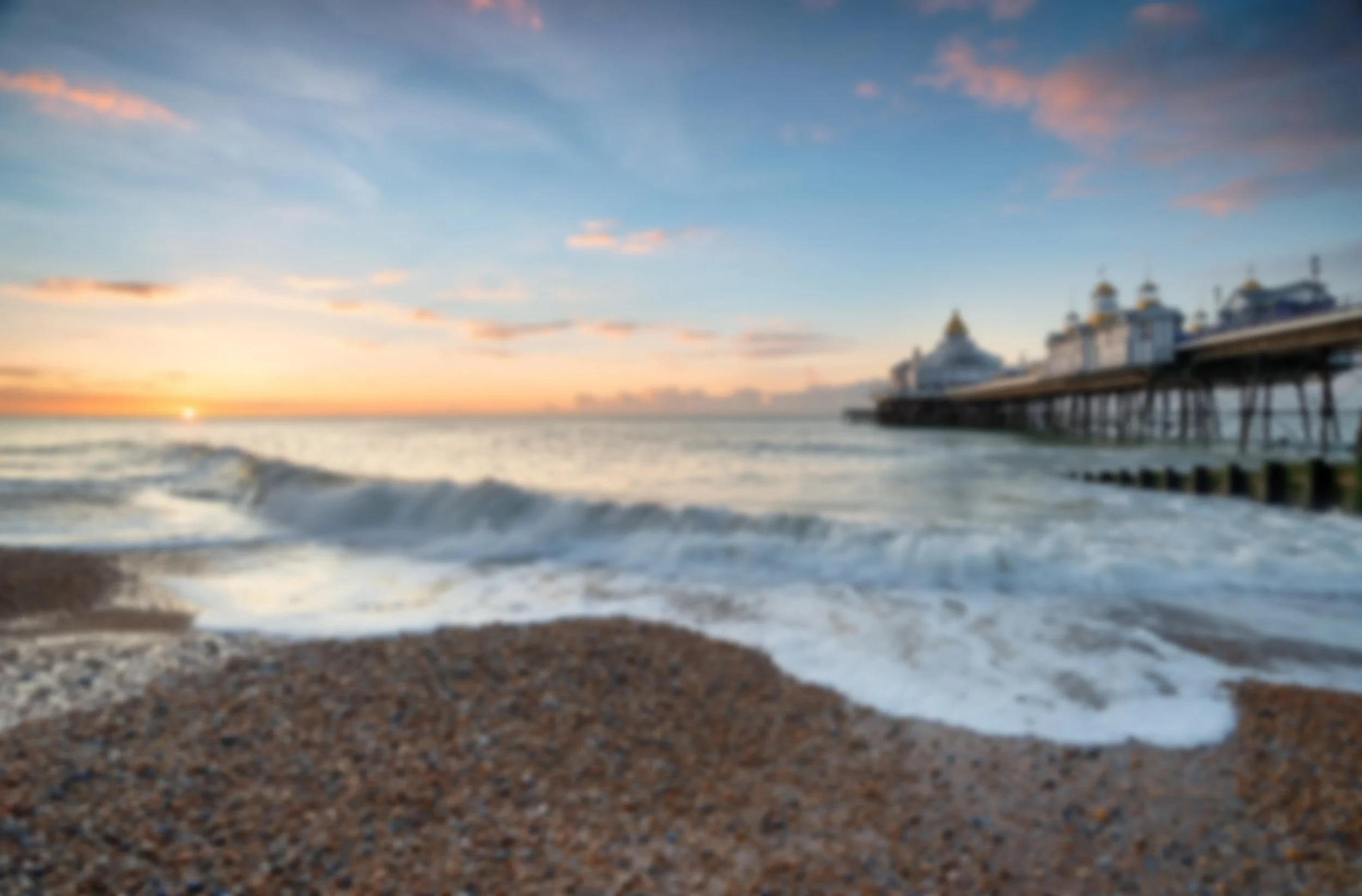 Blurred view of a beach with waves washing onto the sandy shore, a pier extending into the water, and a sunset in the background with blue sky and clouds.