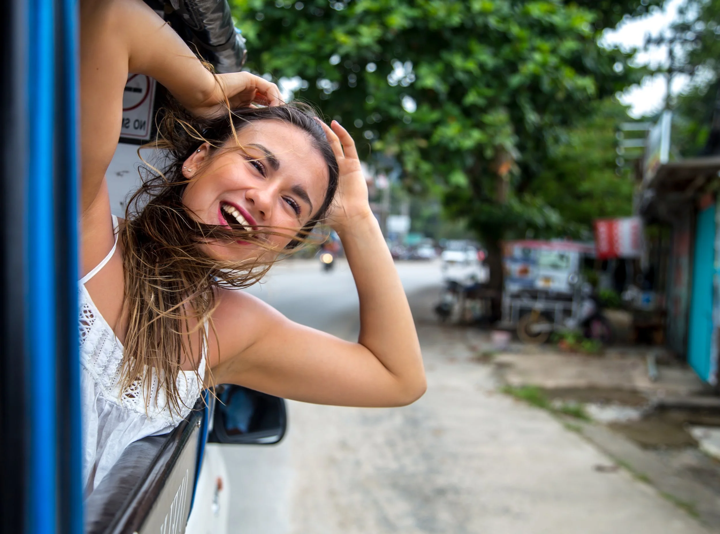 A woman with windblown hair leaning out of a vehicle window, smiling and laughing, with a city street and trees in the background.