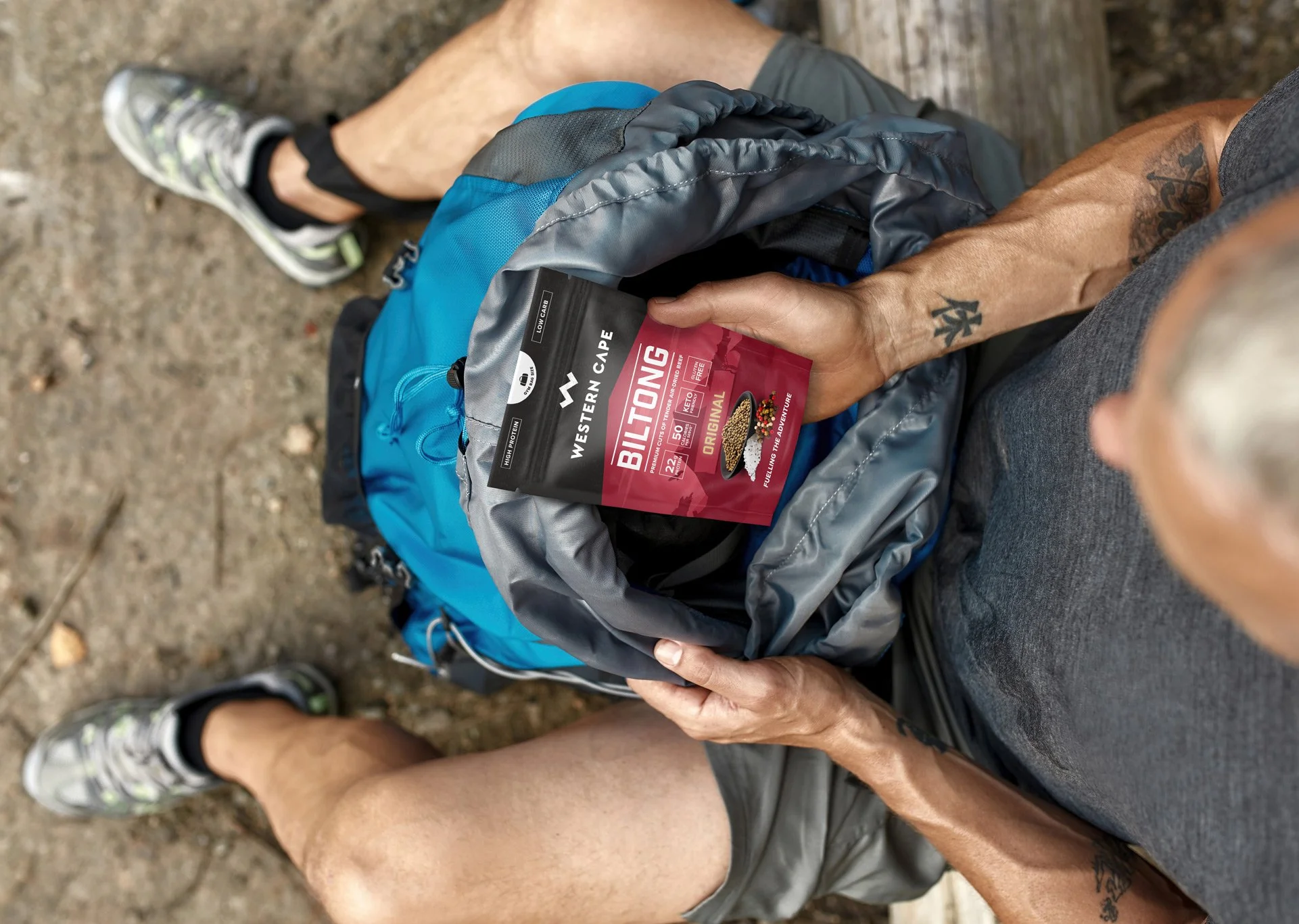 A person sitting outdoors, holding a pack of Western Cape biltong, with a backpack on their lap.
