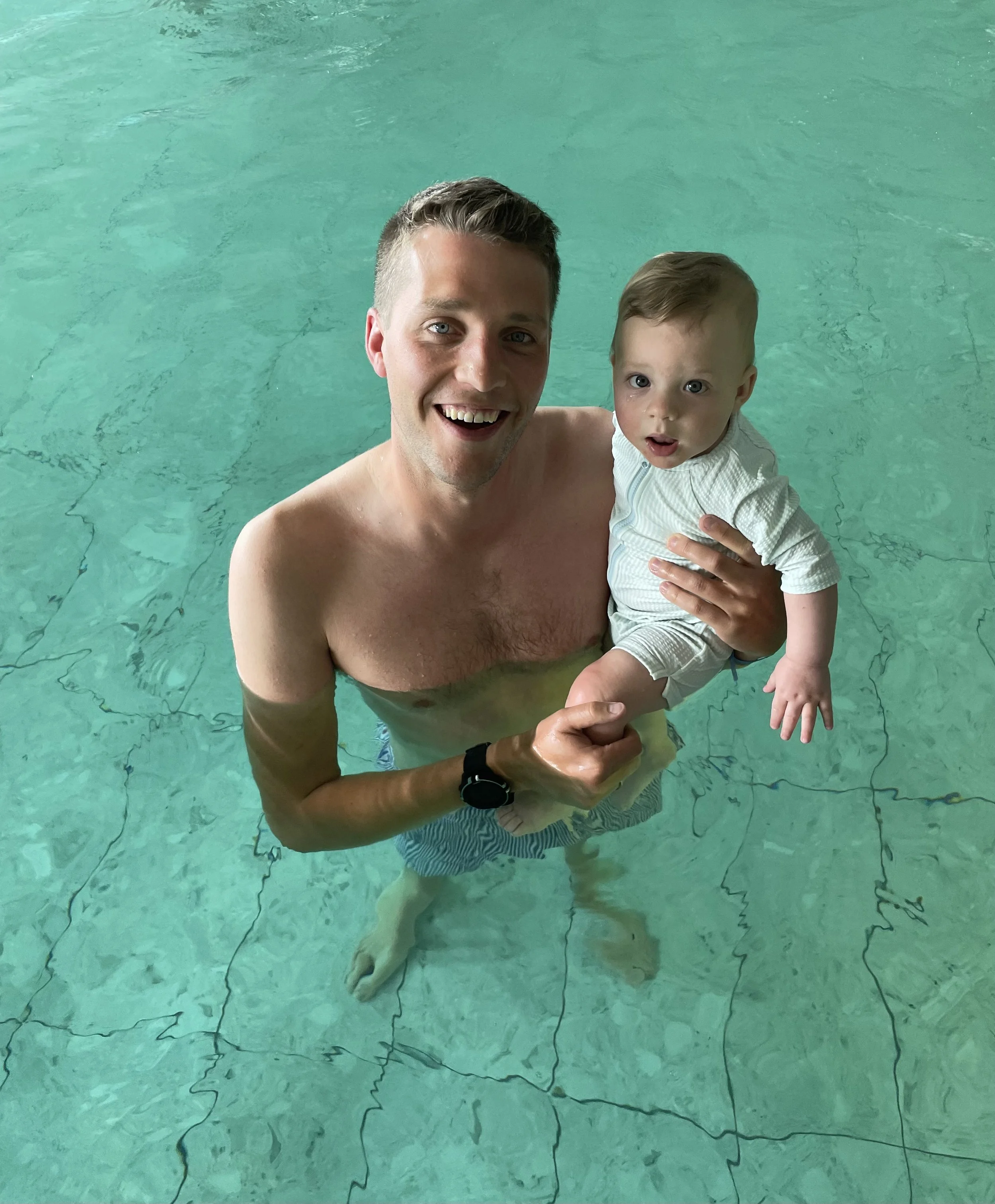 A smiling swim teacher and a young child in a private indoor swimming pool, with the man teaching the child to swim in the water.