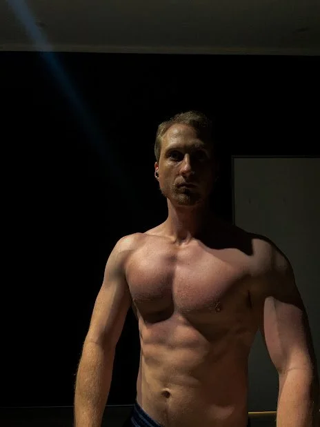 A shirtless man standing in a dimly lit room, taking a mirror selfie.