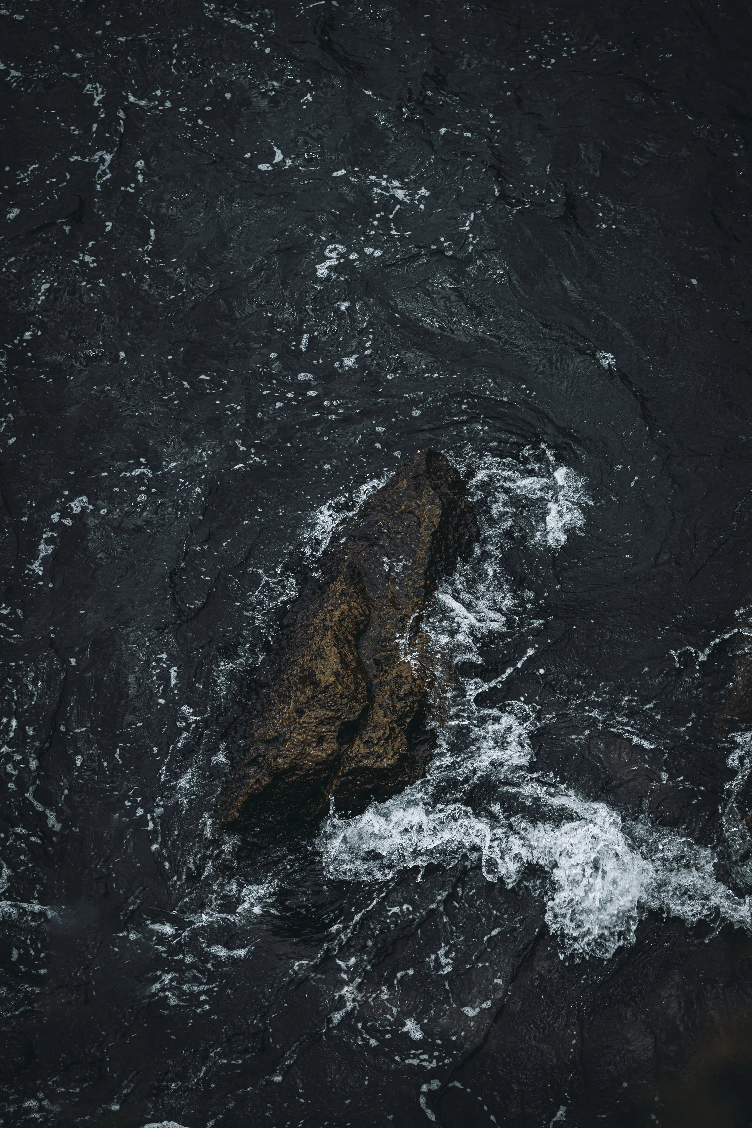Dark water with a large, jagged rock in the center, surrounded by white foam and small ripples.