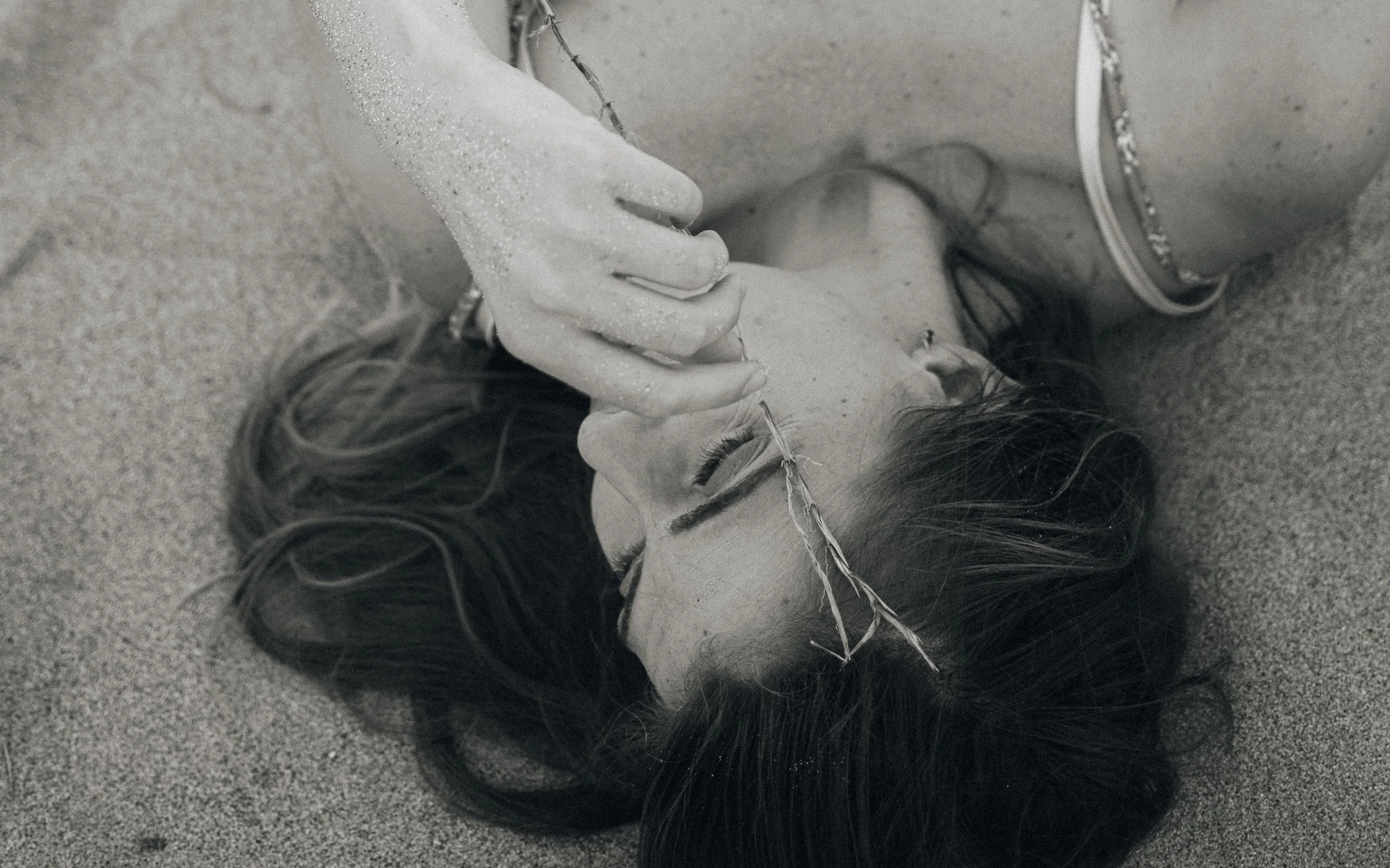 A woman lying on the sand with her eyes closed, wearing a crown made of string and a bracelet on her ankle. She is holding the string crown with her hand and appears to be outdoors.