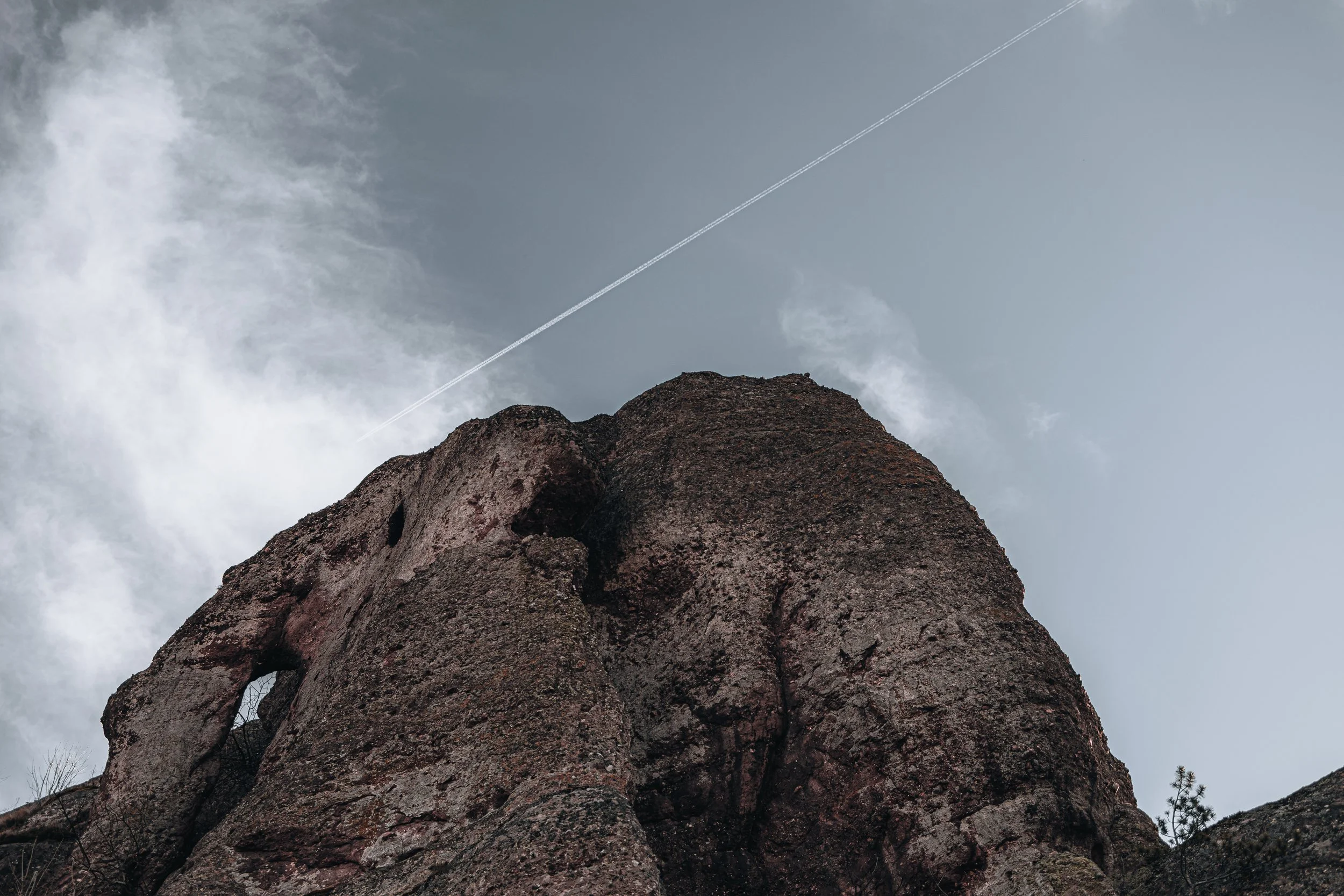 A large rocky formation with a natural arch set against a cloudy sky with a contrail.