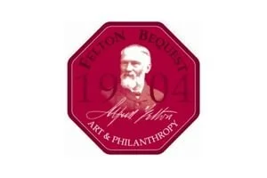 A maroon octagonal badge with a portrait of a man with a beard and mustache at the center. Around the portrait, it says "LUXURY BEAUX ARTS ART & PHILANTHROPY" and the year "1904." There is a signature beneath the portrait.