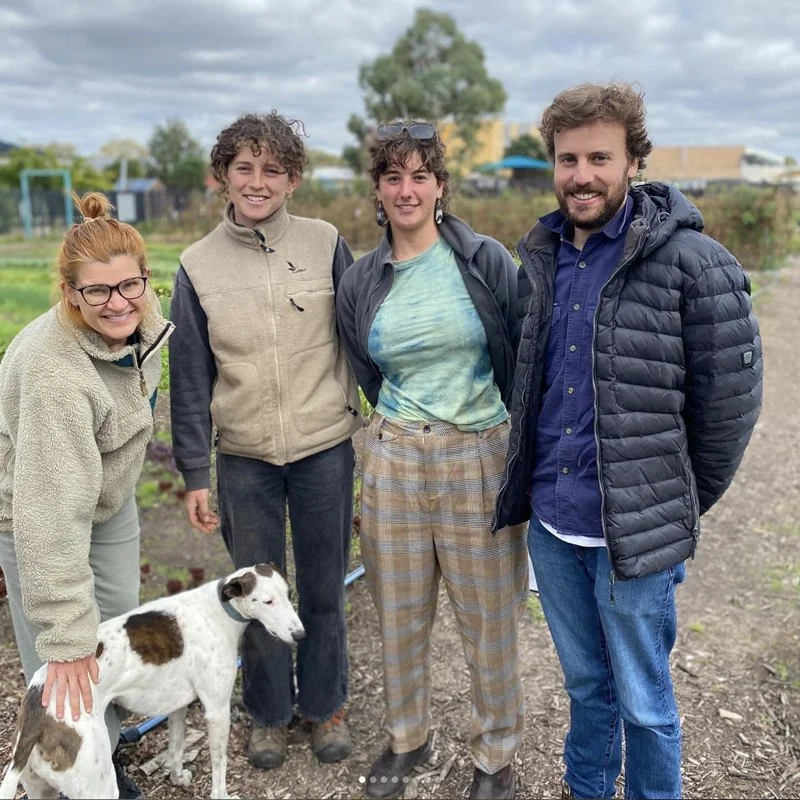 Four people standing outdoors in a garden or farm, with a dog in front. They are smiling, dressed casually, and the weather appears overcast.