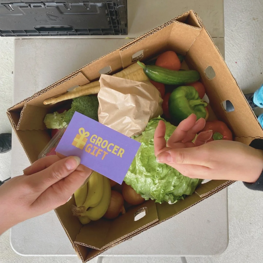 A person holding a card that says 'GROCER GIFT' above a box of fresh vegetables, including lettuce, green bell pepper, zucchini, bananas, broccoli, carrots, and an onion.