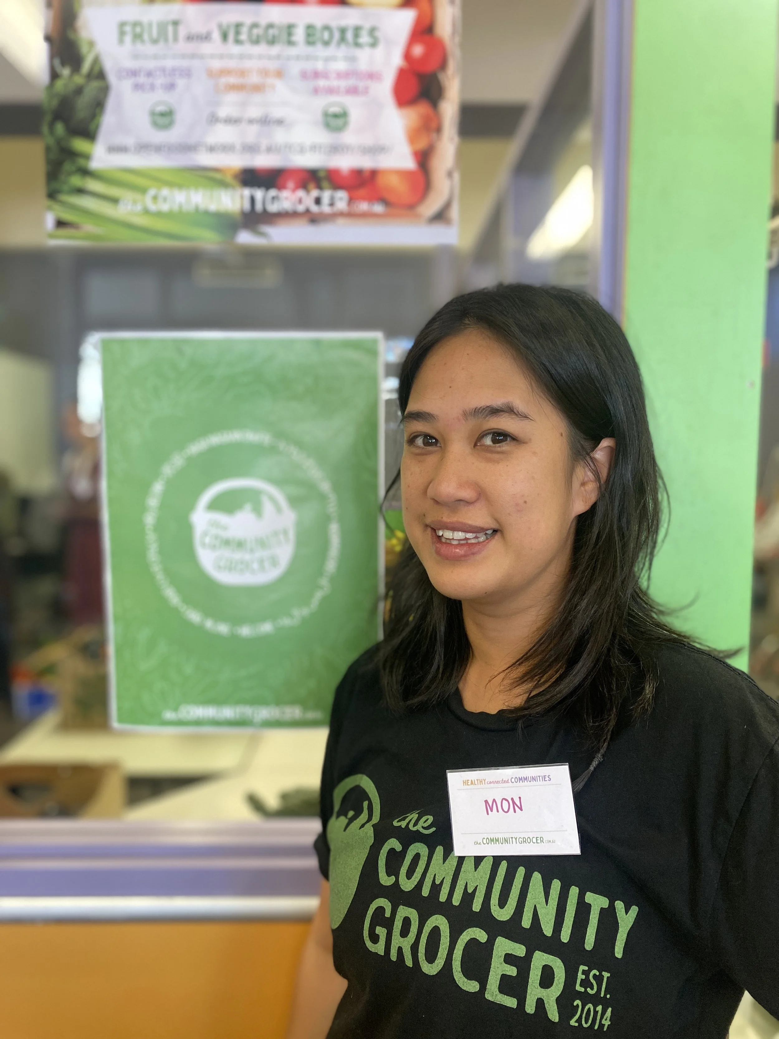 A woman wearing a black shirt with green text that says "the community grocer" and a name tag that says "Mon" standing in front of a sign for The Community Grocer. The sign has a green background with the store's logo, a shopping basket with a leaf, and other promotional material about fruit and vegetable boxes.