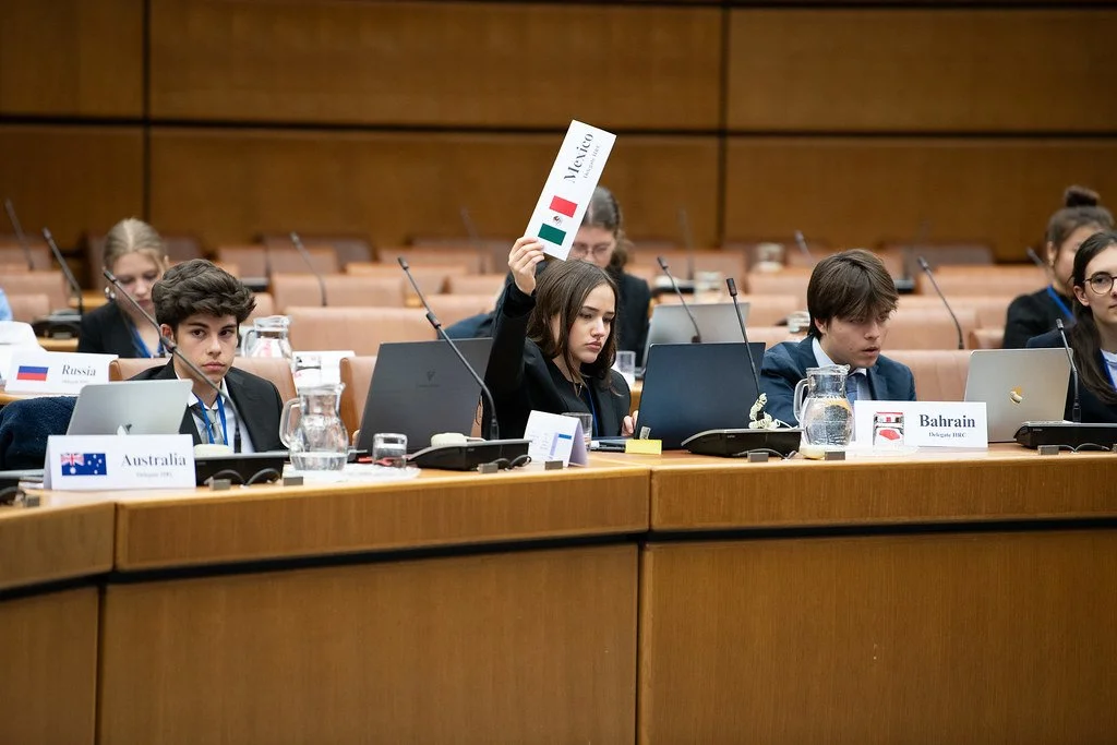 Young delegates at an international conference, with a woman from Italy raising a hand holding a small Italian flag. Other delegates have nameplates for Russia, Australia, Bahrain, and Italy visible on the table.
