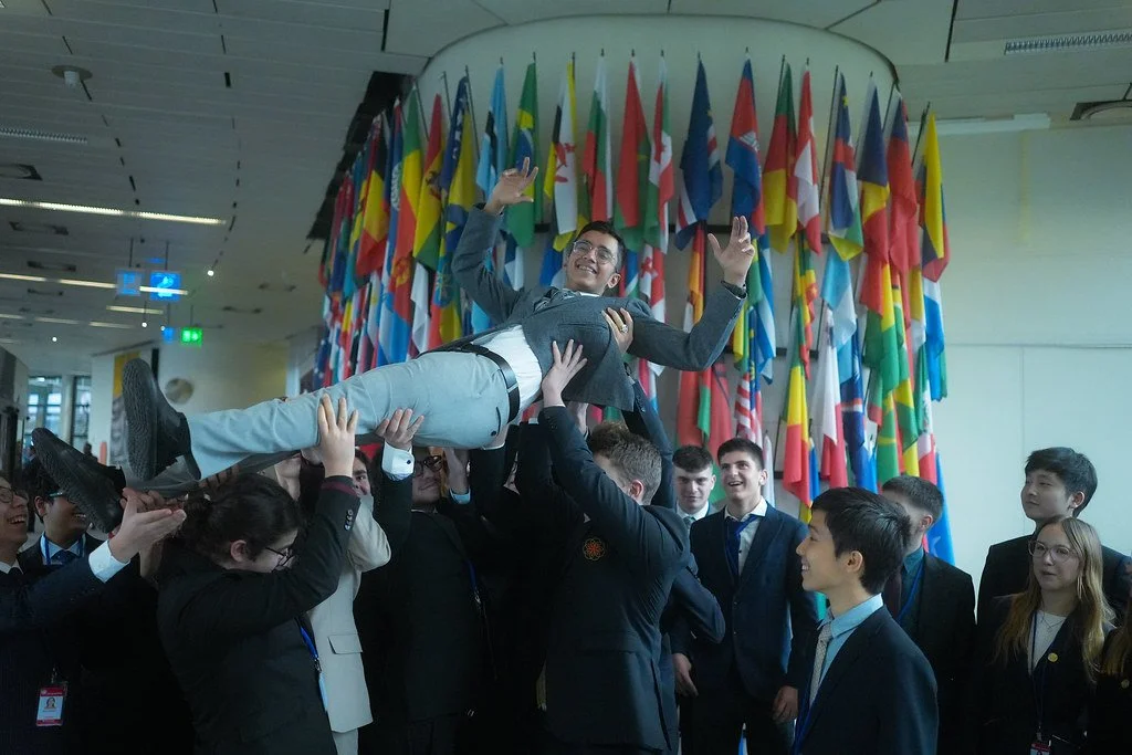 A group of people in business attire lifting a man into the air at an international event, with many national flags in the background.