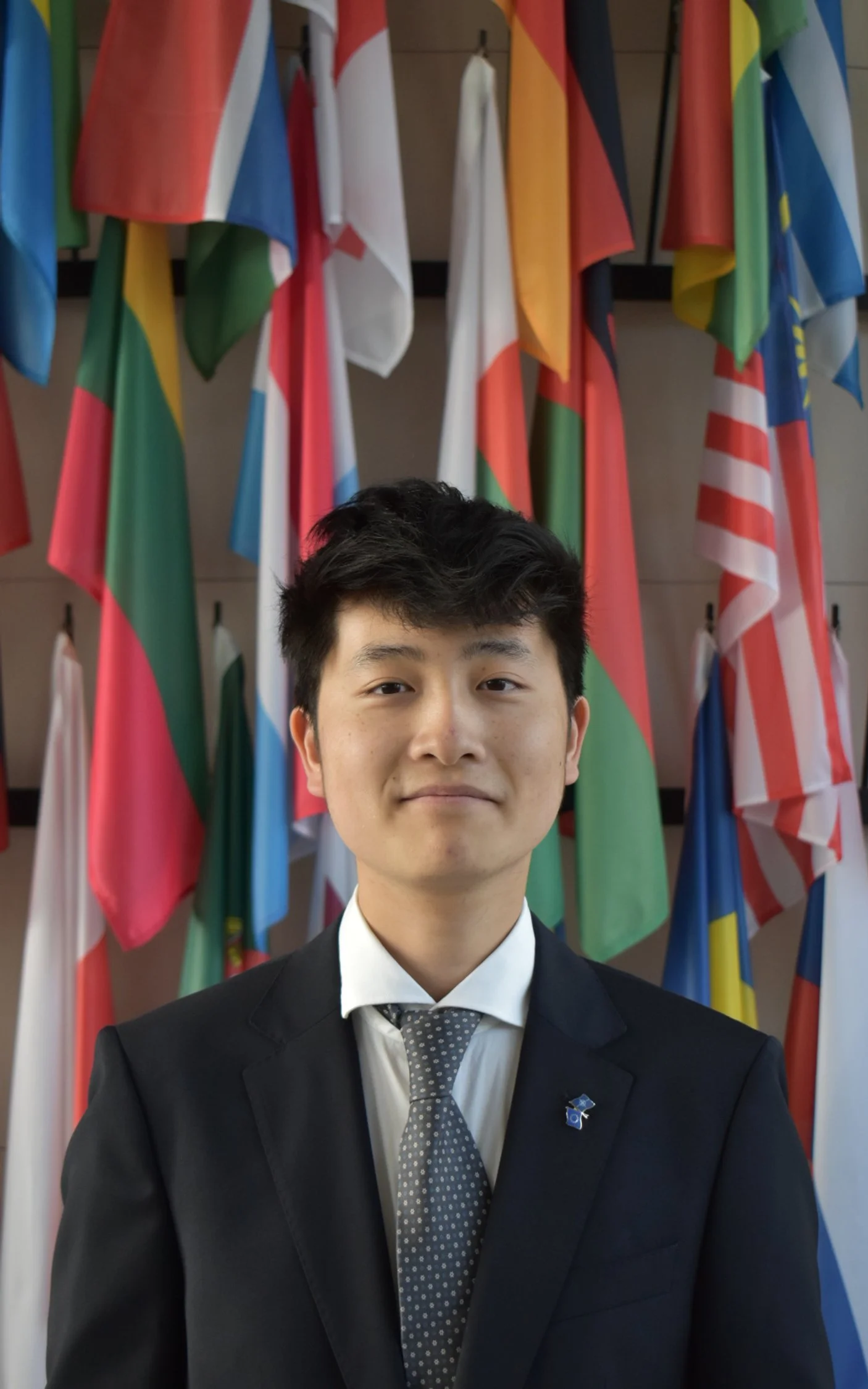 A young man in a black suit, white shirt, and blue patterned tie, smiling in front of a plain, light-colored background.