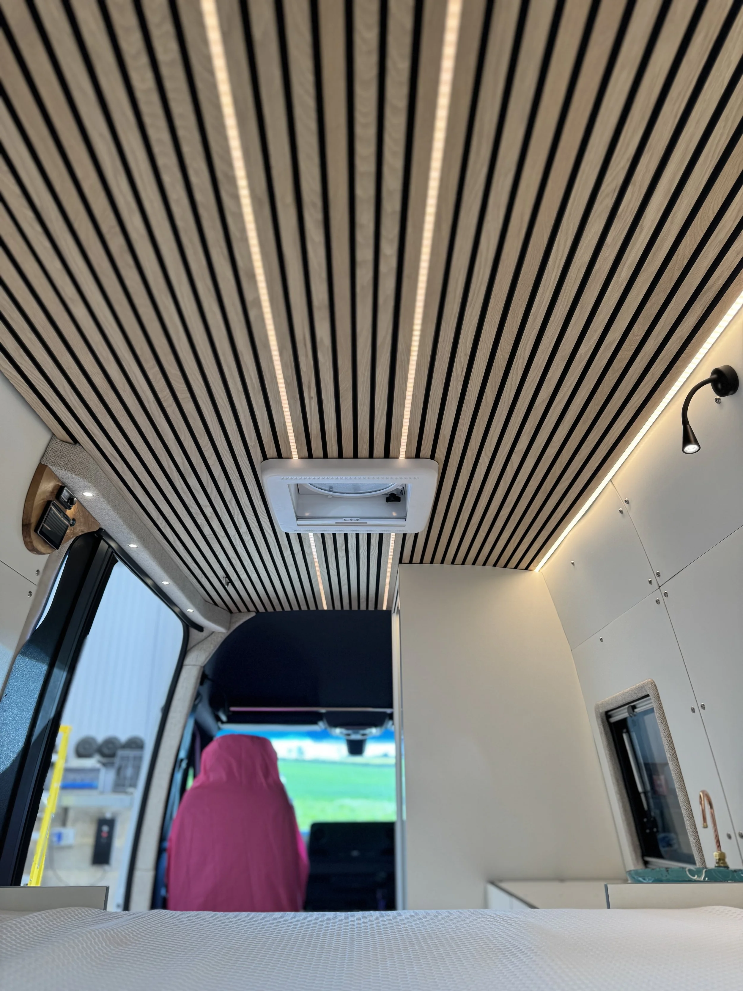 Interior of a modern vehicle with wooden slatted ceiling, a mounted air conditioning unit, and a pink seat cover on the driver seat visible through the front window.