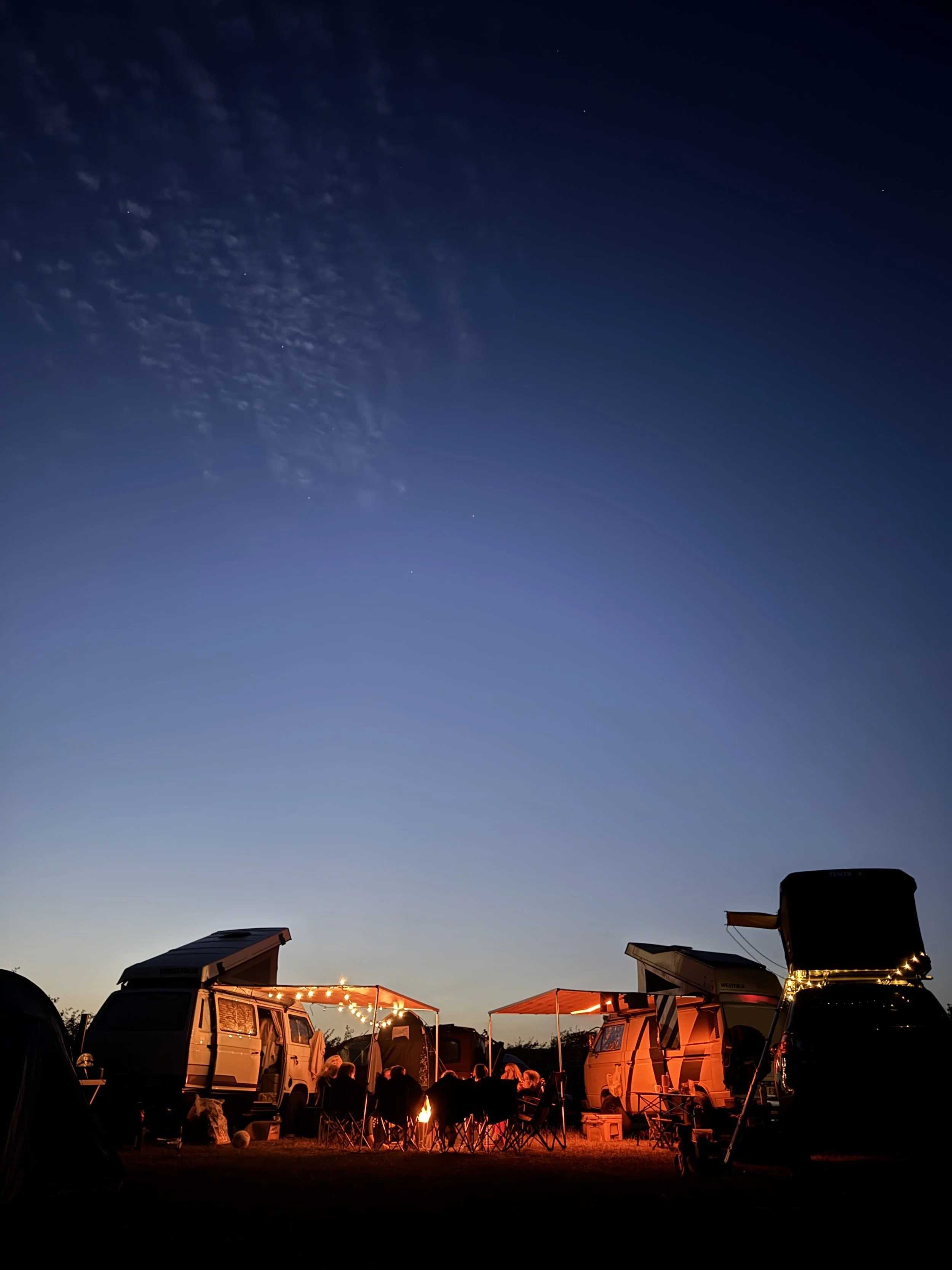 Campground with several pop-up campers and a group of people sitting around a campfire under a darkening sky with stars and some clouds.