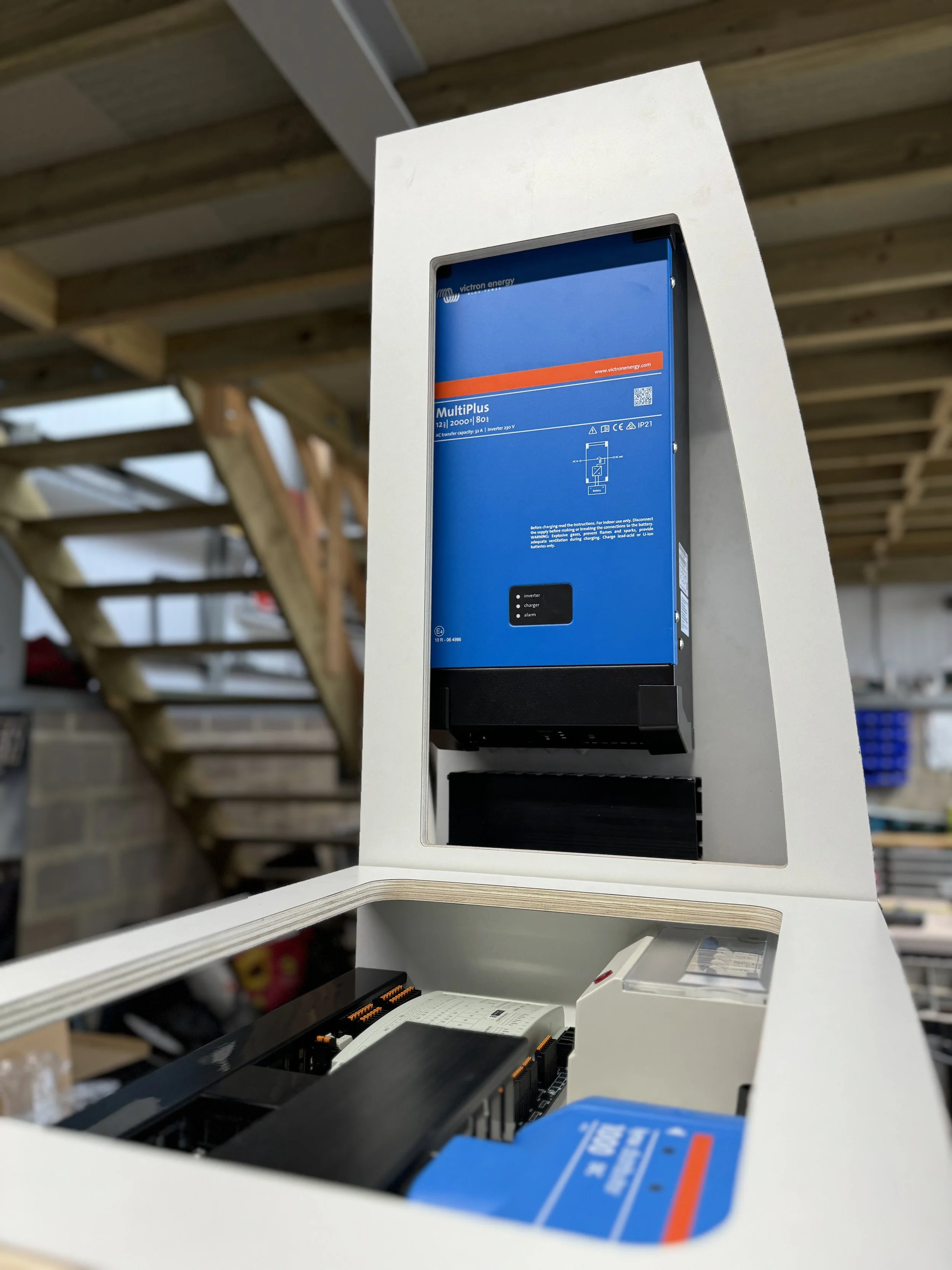 Close-up of an open electrical panel with a blue Victron energy MultiPlus inverter/charger inside, located in a workshop with wooden ceiling beams.
