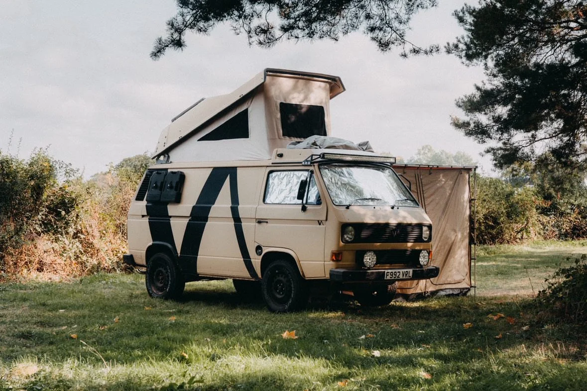 A beige camper van with a pop-up roof, parked on grass in a wooded area.