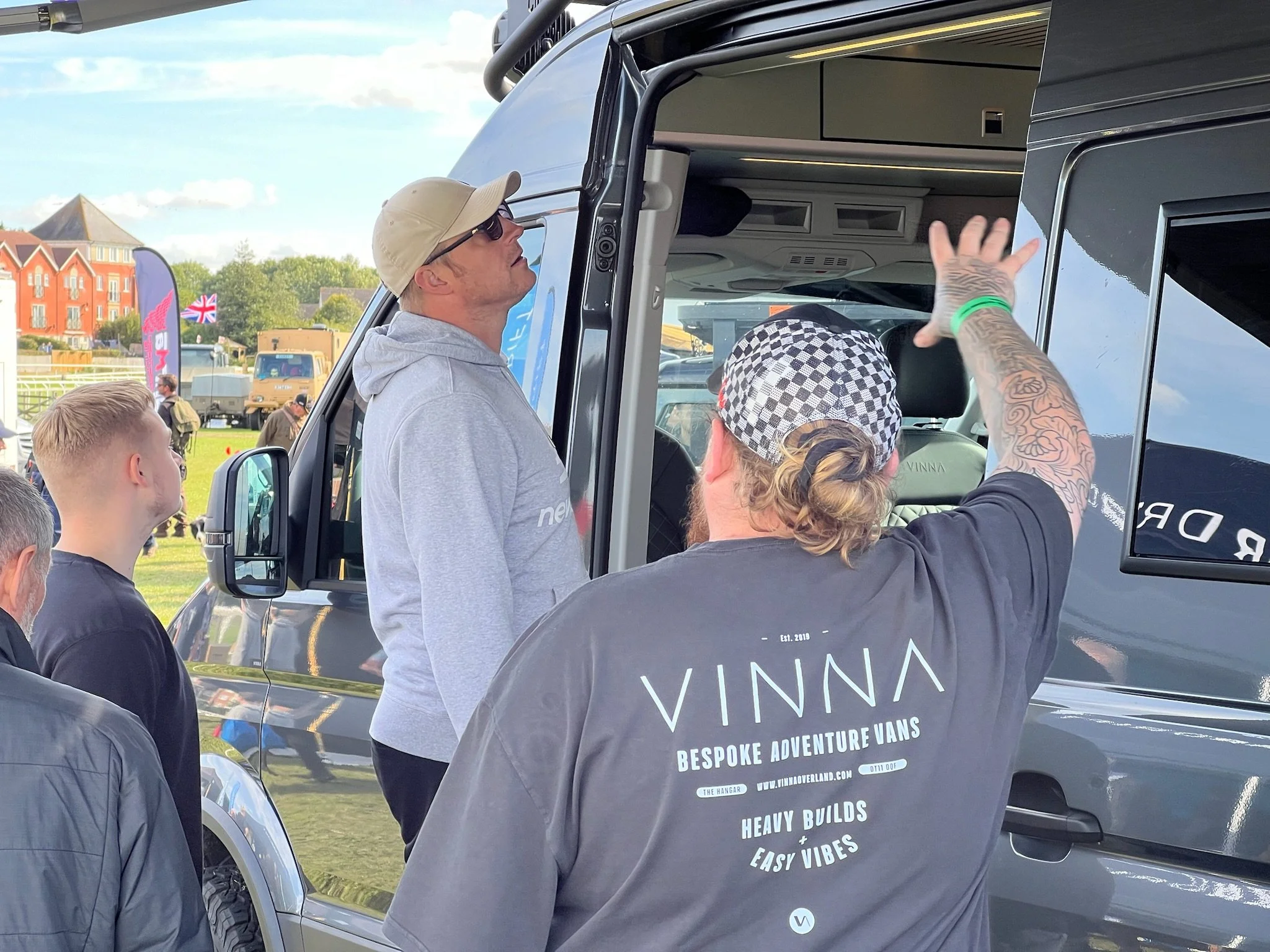 Men gathered around a large van, with one man in a gray hoodie and sunglasses looking up at the van, and another man with long hair, tattoos, and a checkered cap gesturing towards it. In the background, there are buildings, flags, and a grassy area.