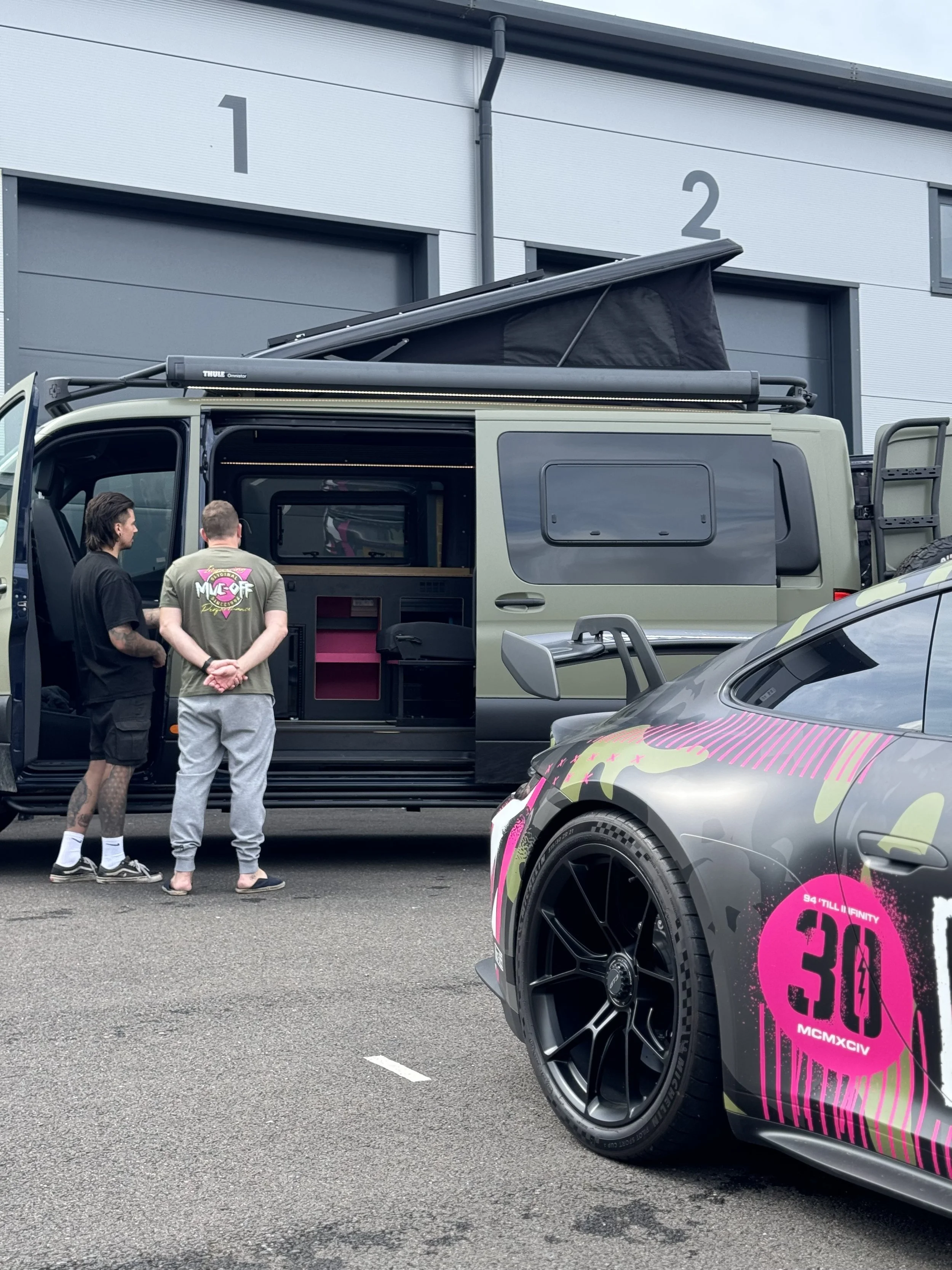 Two men are standing outside a 4x4 Sprinter van with a pop-up roof, in front of a gray warehouse with numbered doors 1 and 2. A Porsche 911 with colorful graphics is parked nearby.
