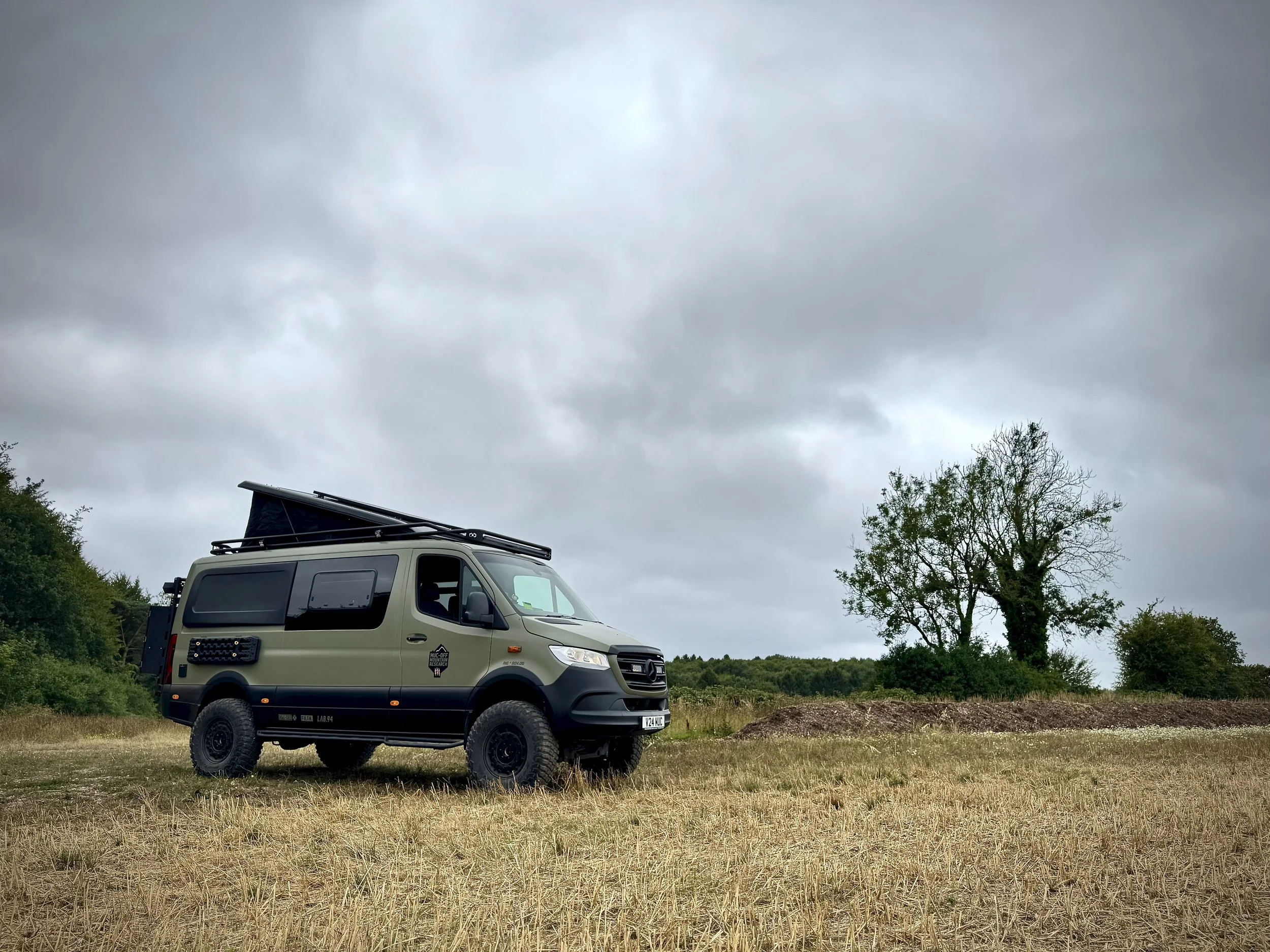 A camouflaged off-road camper van parked on a grassy field under a cloudy sky, with a tree in the background.