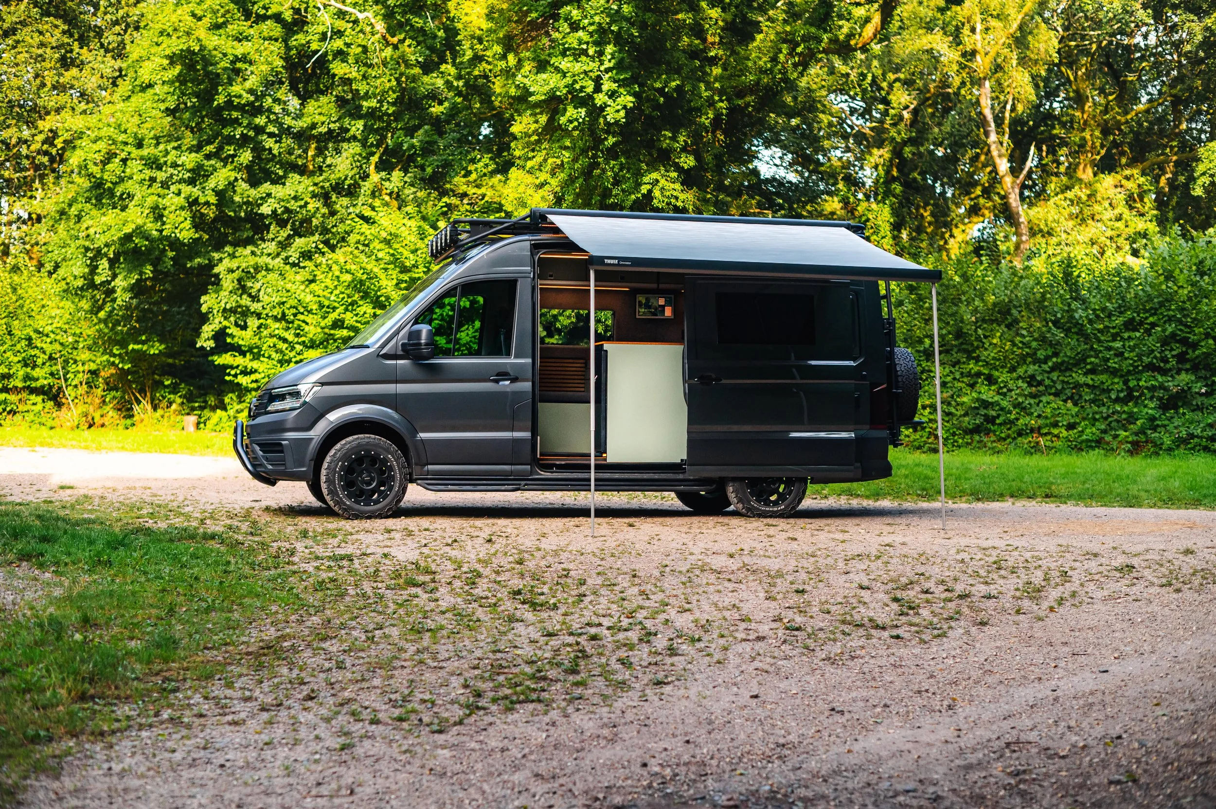 A black camper van with an extended awning parked on a gravel surface in a lush green forest.