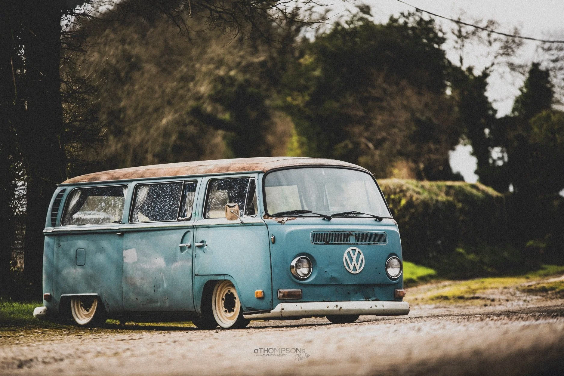 A vintage blue Volkswagen bus with a weathered appearance parked on a dirt path surrounded by trees.
