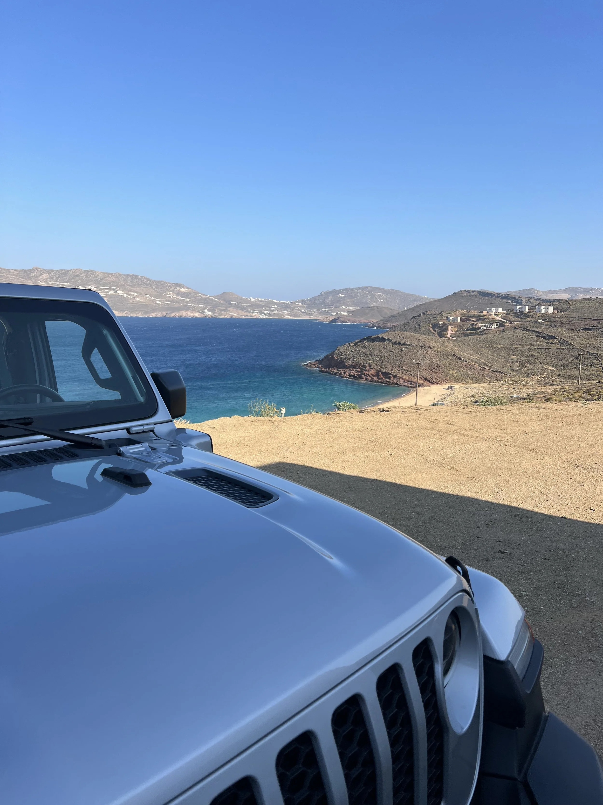 Close-up of the front part of a silver Jeep SUV parked on a dirt road overlooking a coastal landscape with blue ocean, hills, and a clear sky.