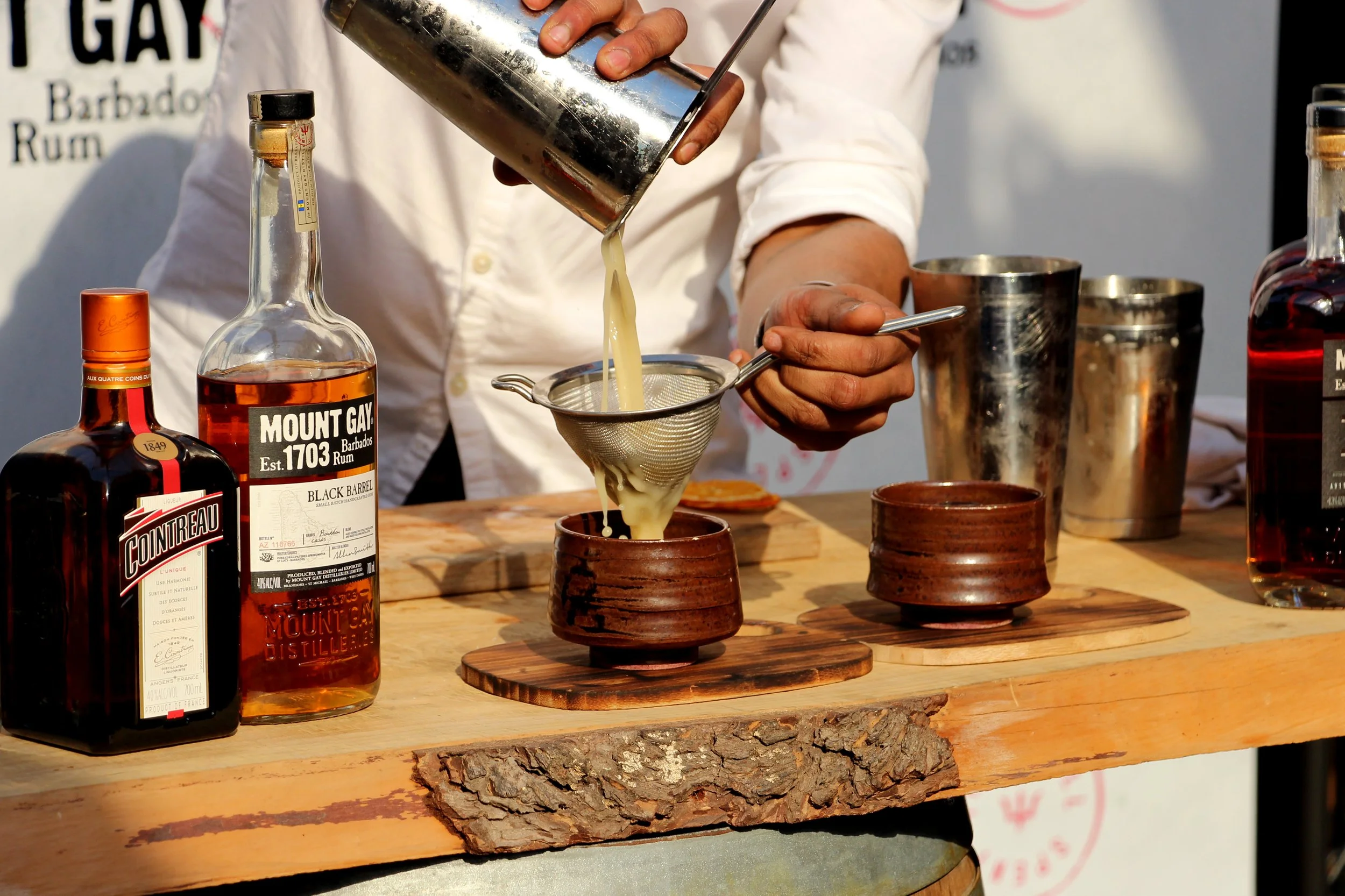 A bartender pours a liquid through a strainer into a small brown cup filled with yellow drink, with bottles of rum and metal drinkware on the wooden bar.