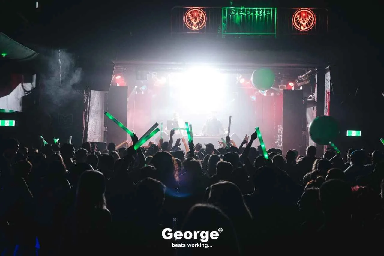 Crowd at a nightclub or concert with glow sticks, facing a stage with DJ equipment, bright LED screen, and promotional banners, during a nighttime event.