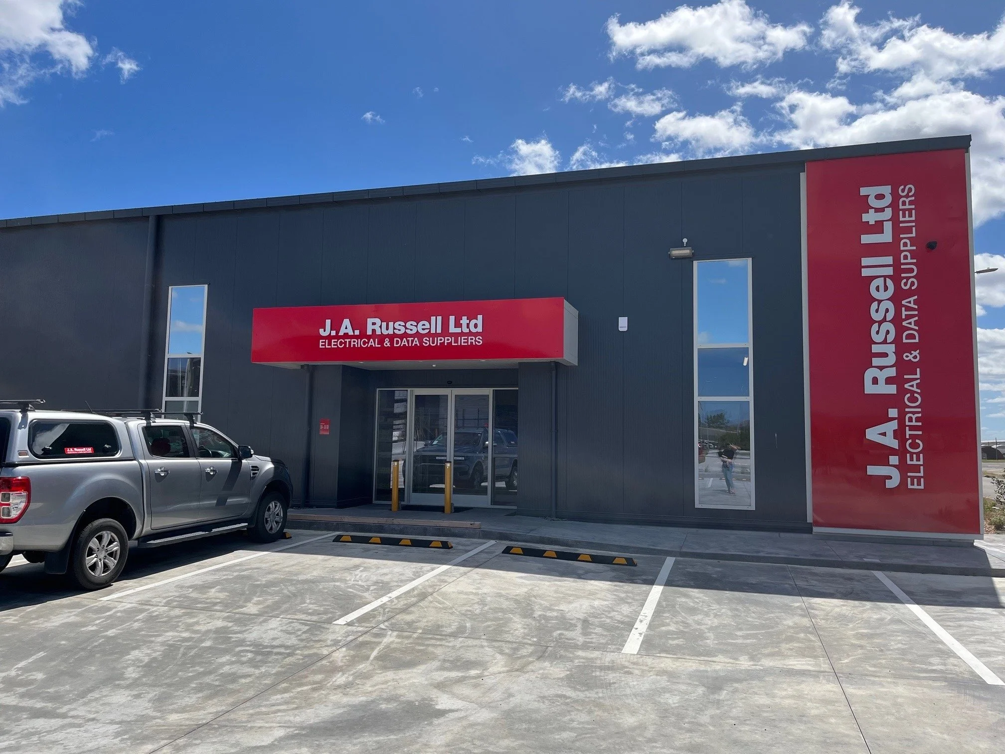 Exterior view of J. A. Russell Ltd store with parking lot in front, a grey pickup truck, blue sky with clouds, and store signage indicating electrical and data supplies.