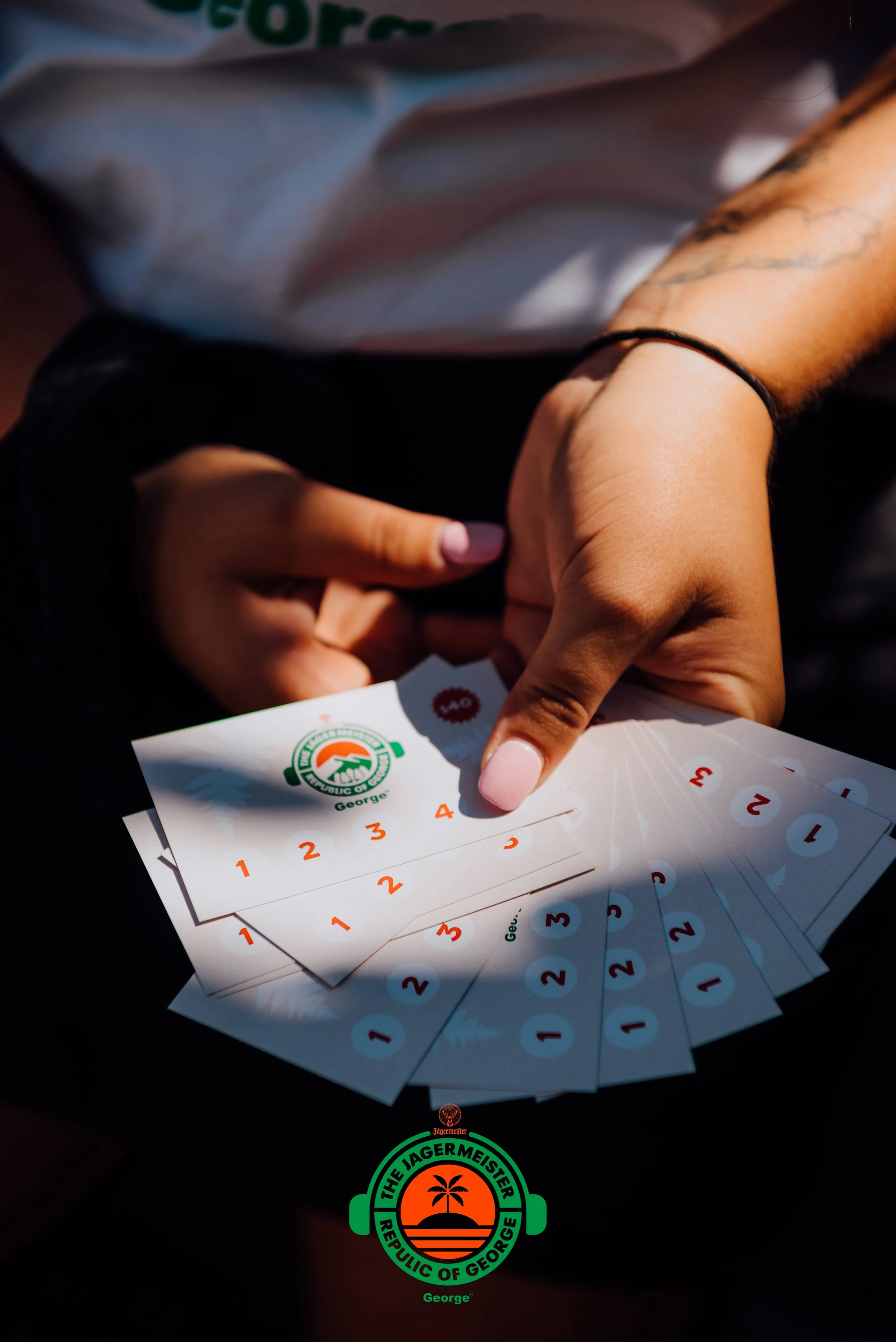Person holding multiple cards with the logo of the Jägermeister Republic of George, showing a landscape with a palm tree and sunset.