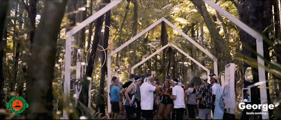 A group of people gathering outdoors under a white geometric arch structure in a wooded area.