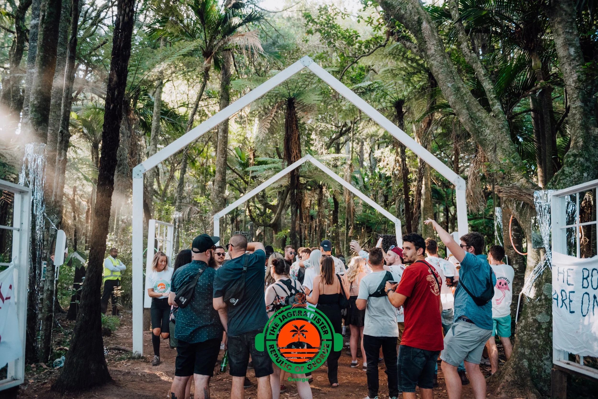 A large group of people gathered outdoors in a forested area for a party or event, with wooden arch frames decorated with string lights and signs, some people are talking and using their phones, and there is a security guard visible in the background