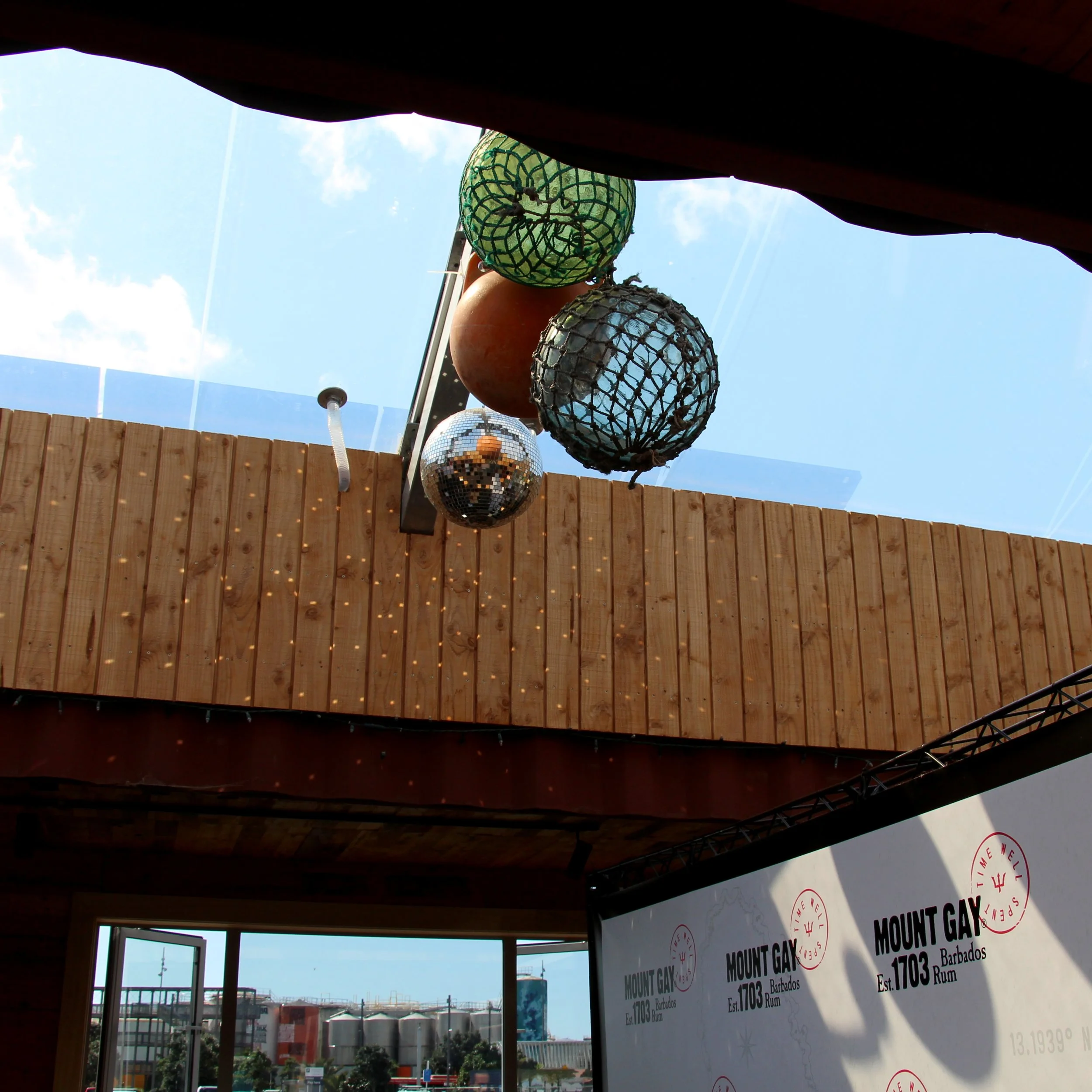 View of hanging decorative lanterns and a disco ball from below, with wooden wall and blue sky in the background, at a bar or restaurant in Barbados.