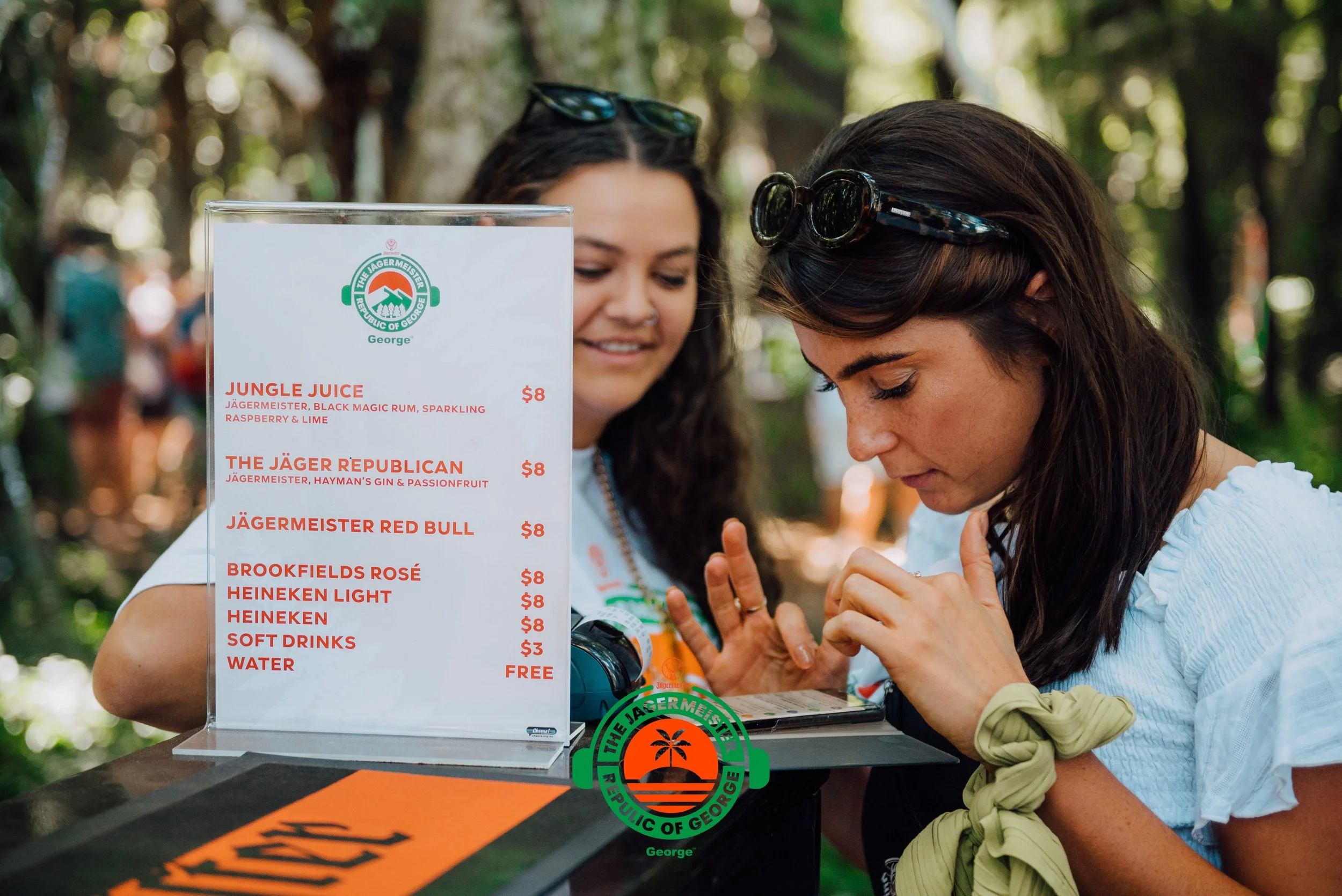 Two women at an outdoor bar looking at a menu, surrounded by a wooded area. The menu features various drinks and prices, with a green and orange logo of the Jagermeister brand.