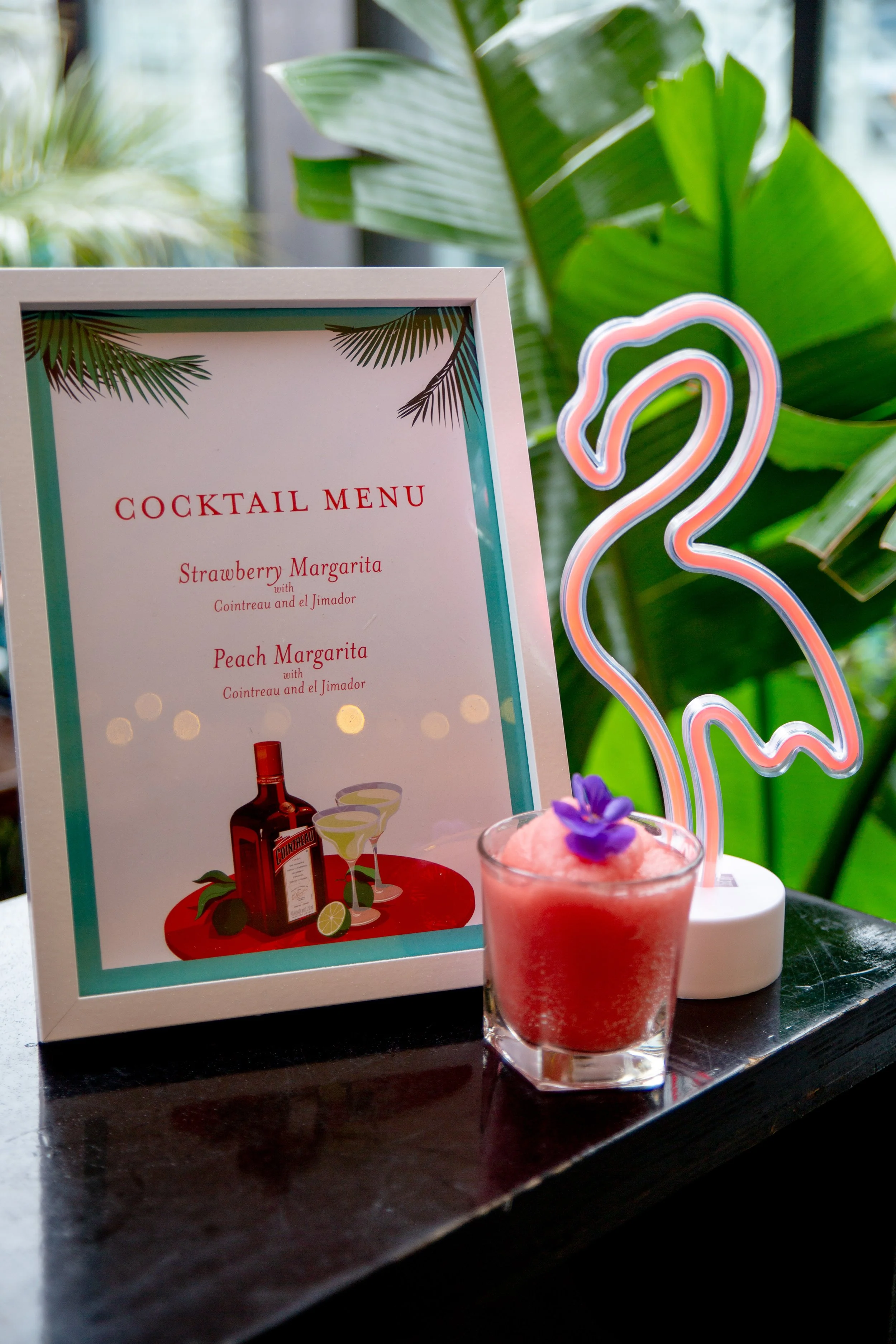 A pink cocktail garnished with a purple edible flower, a neon flamingo light, a cocktail menu with tropical leaves, and greenery in the background.