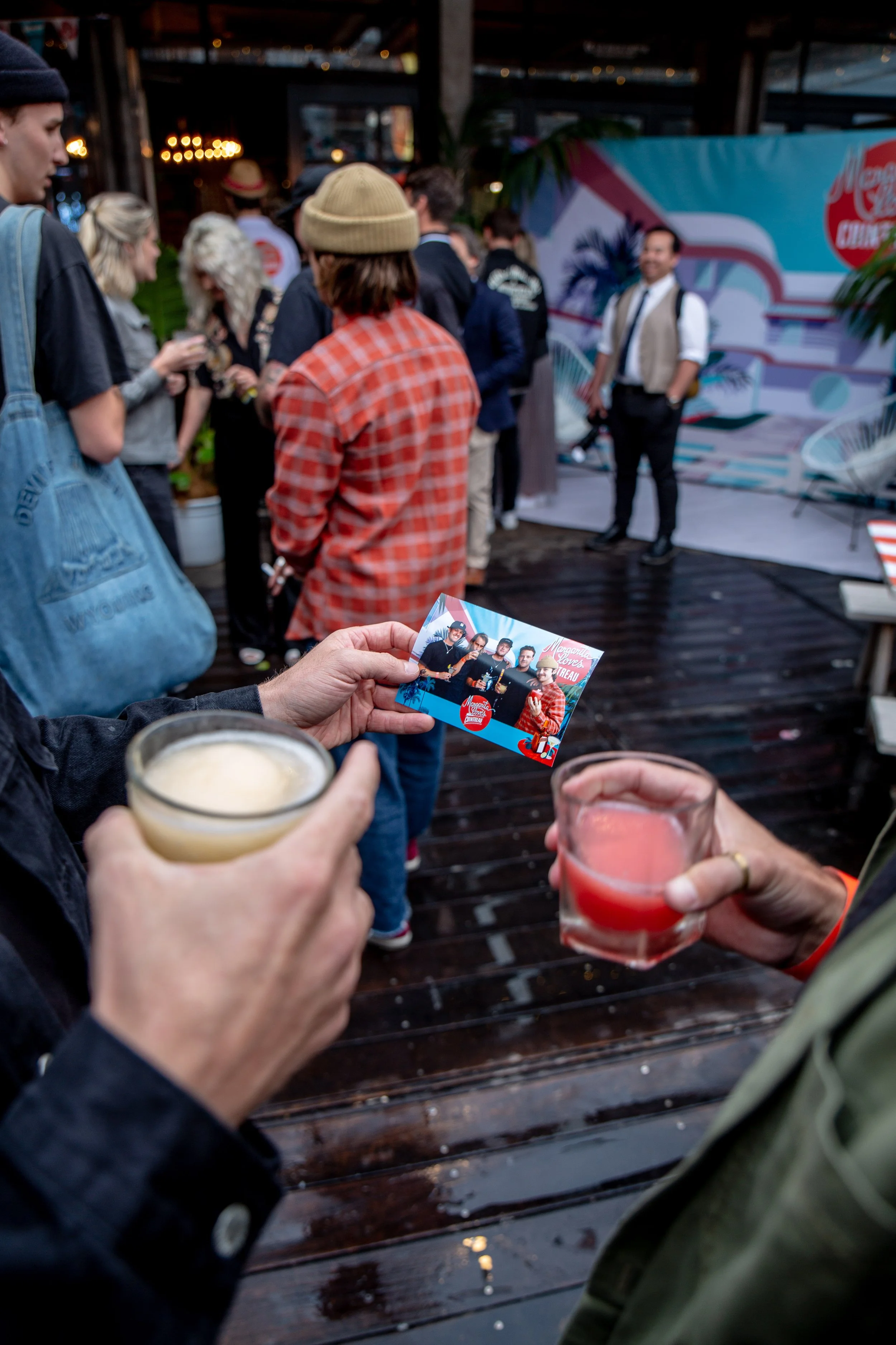 People at a social gathering holding drinks, with a person in the foreground holding a photo showing a band or musical group, and a stage with a colorful backdrop in the background.