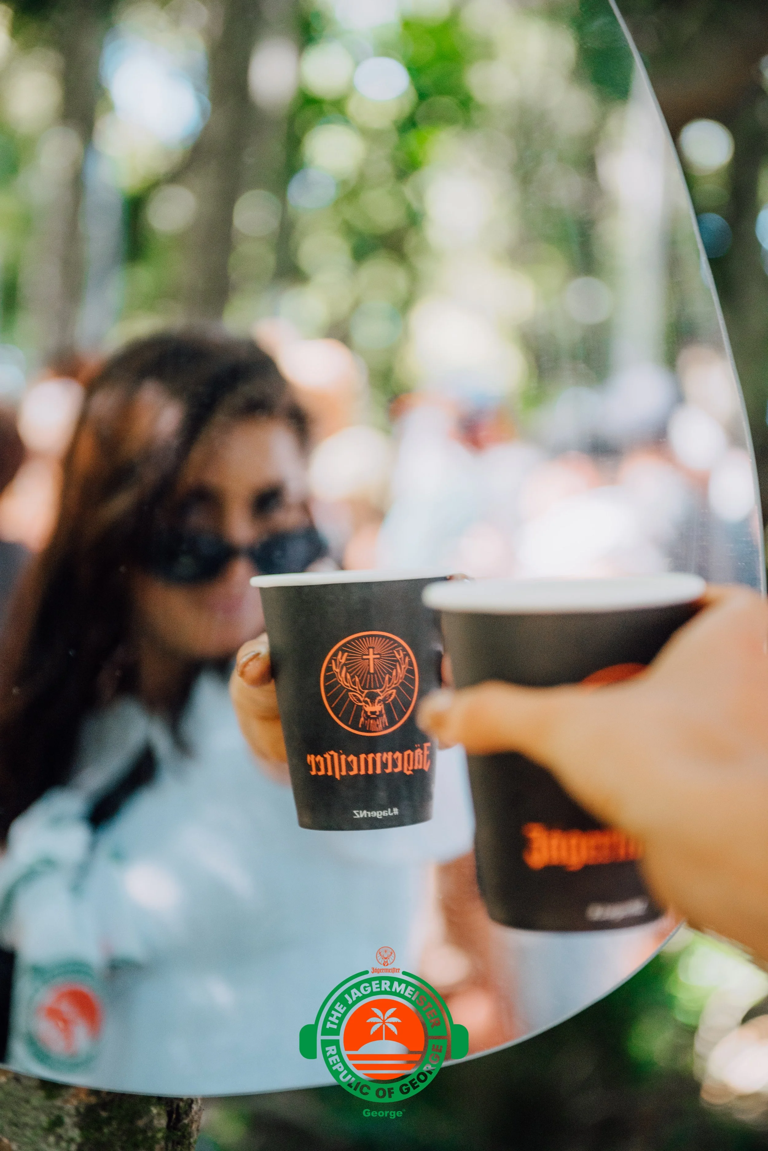 A woman with sunglasses in the background looking at a mirror reflecting two cups of Jägermeister, with a logo at the bottom reading 'The Jägermeister Republic of Georgia, George'.