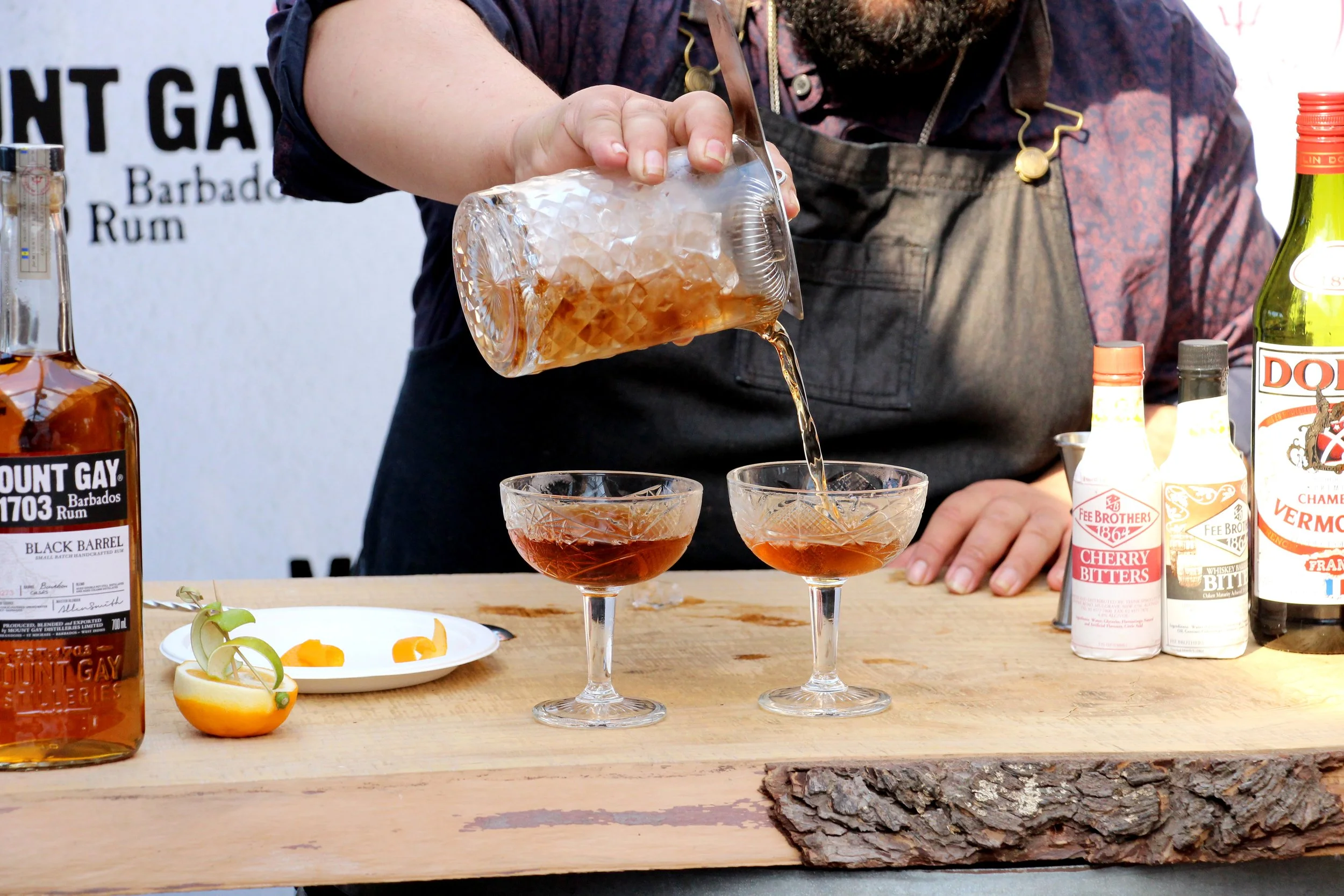 Person pouring a cocktail from a glass pitcher into two coupe glasses on a wooden bar counter, with bottles of alcohol and garnishes in the background.