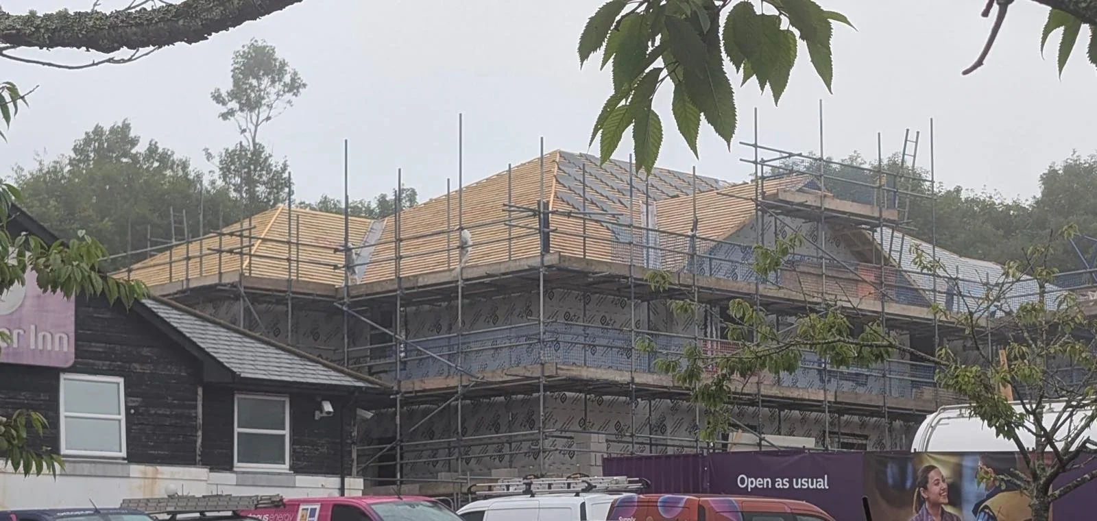 A house under construction with scaffolding around it, a partially completed roof, and construction materials. Trees and construction vehicles are in front.
